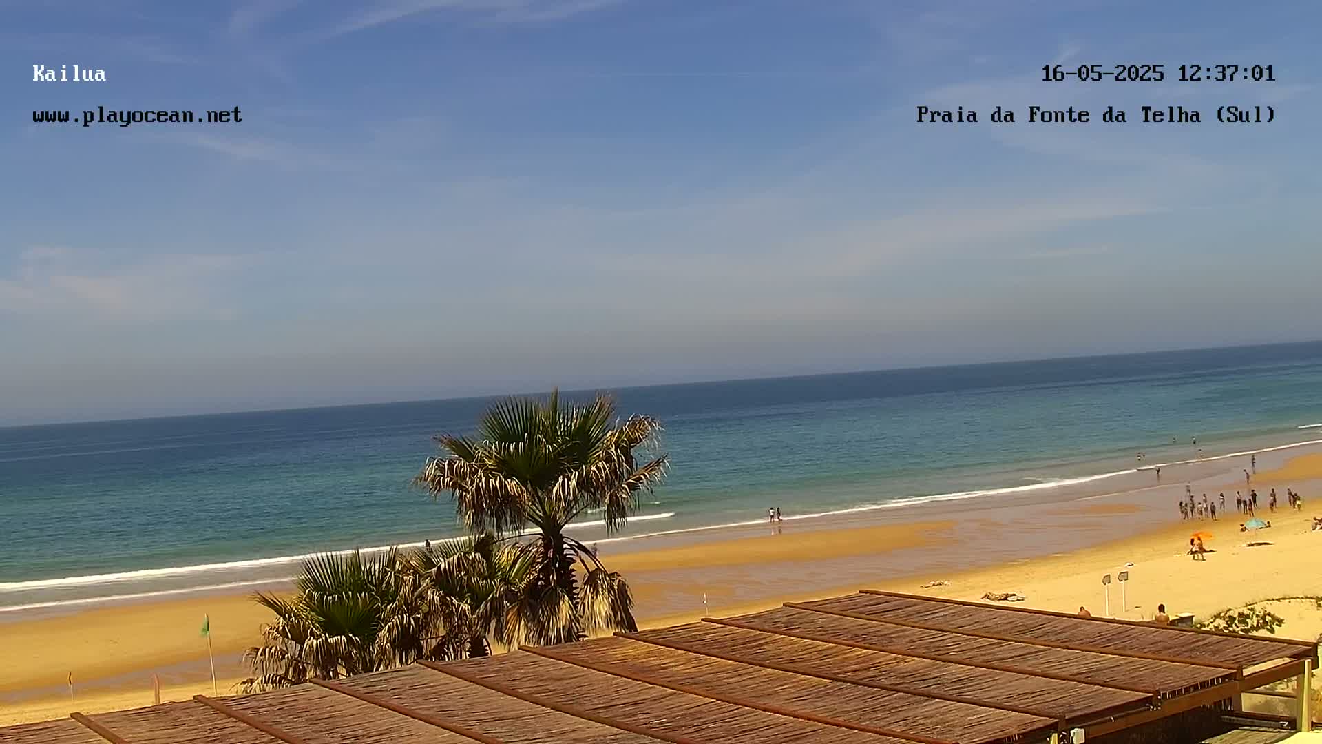 A sandy beach with calm ocean waves and scattered people is viewed from above a thatched roof structure under a clear, sunny sky.