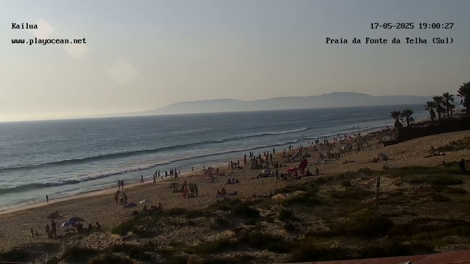 A sandy beach with many people on it is shown, under a clear, calm evening sky with a distant mountain range visible across the ocean.