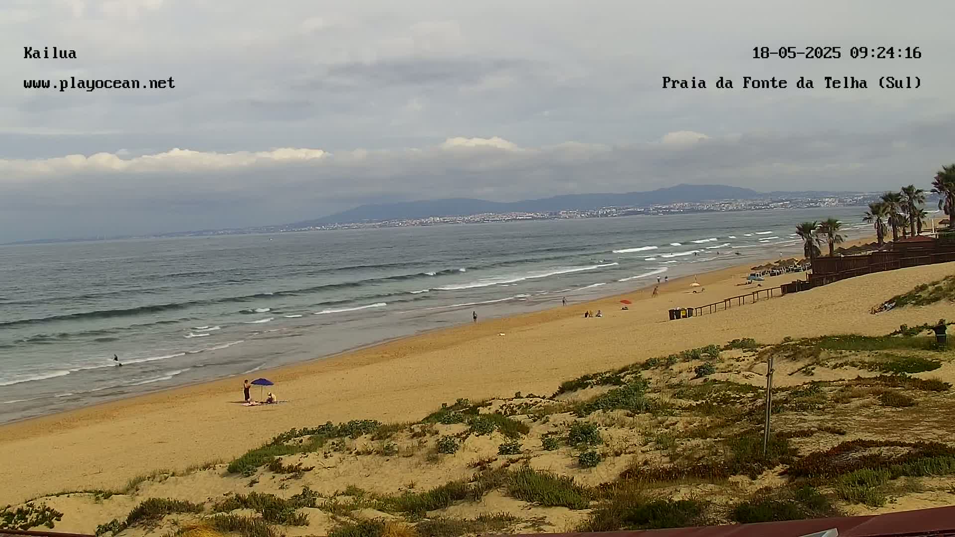A sandy beach with a few people on it, gently rolling waves in the ocean, and a partly cloudy sky, with distant mountains visible on the horizon.
