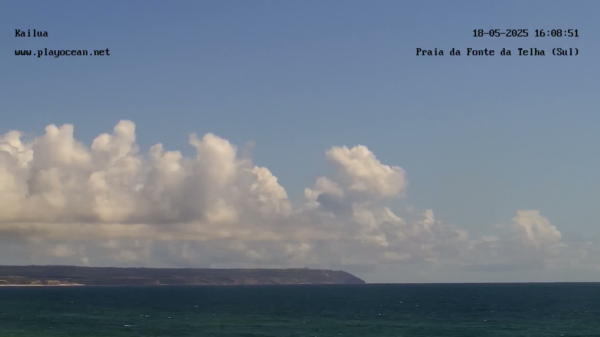 A mostly sunny sky filled with fluffy white cumulus clouds over a dark blue ocean and a distant landmass.