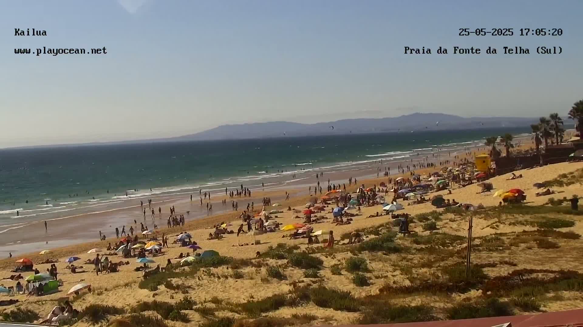 A sandy beach with many people under umbrellas, facing a calm ocean with some kitesurfing in the distance under a clear sky.