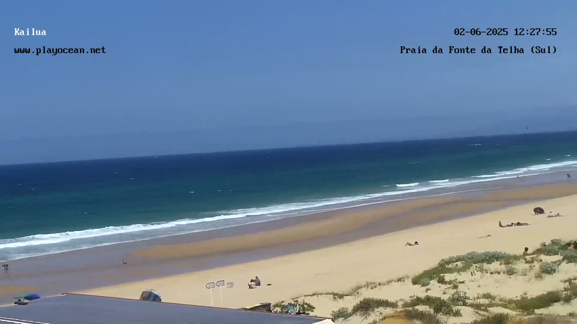 A wide sandy beach with a few people on it slopes down to a calm, teal ocean under a clear blue sky.