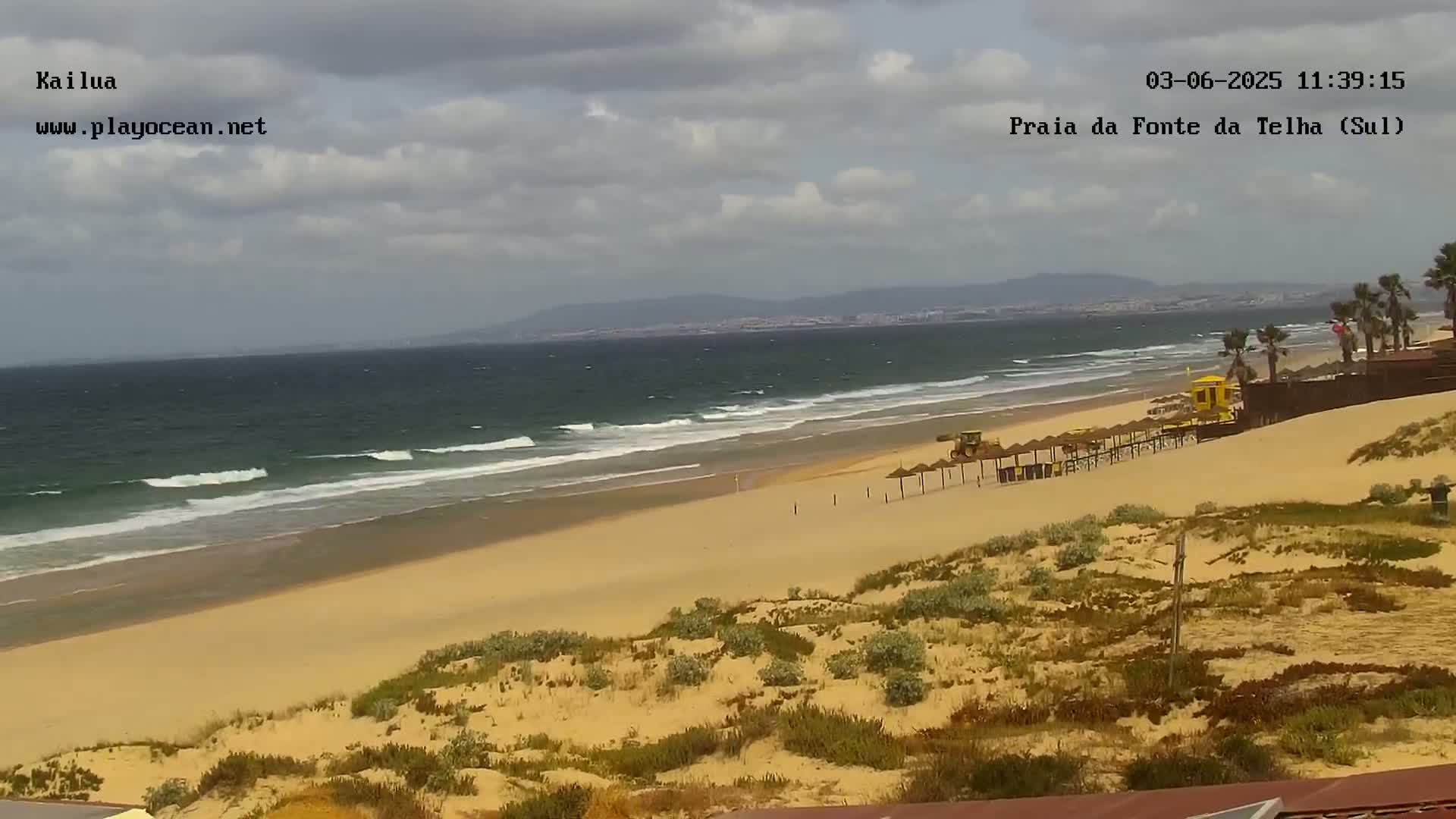 A sandy beach with small waves under a cloudy sky, featuring beach umbrellas and a lifeguard stand, is viewed from a dune overlooking the ocean.