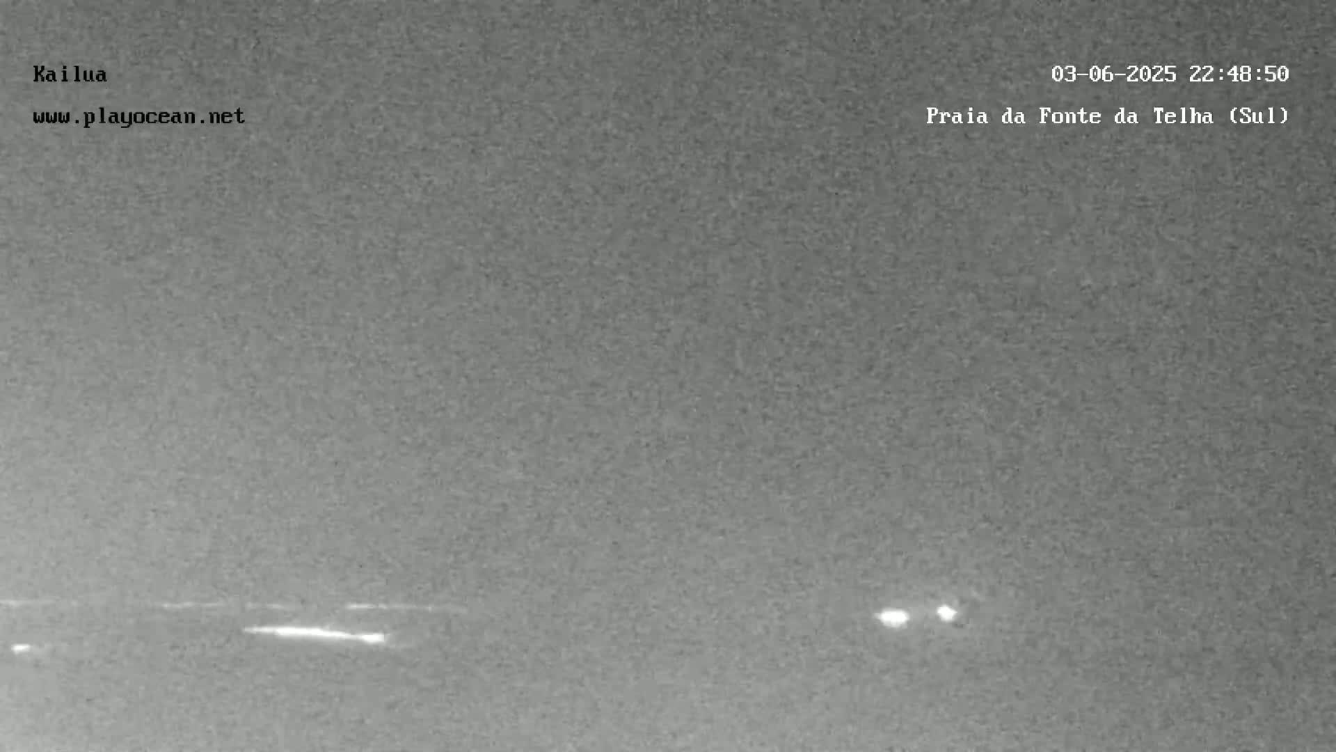 A nighttime, low-light image shows a dark, flat expanse of land or water with a few faint lights visible in the distance.