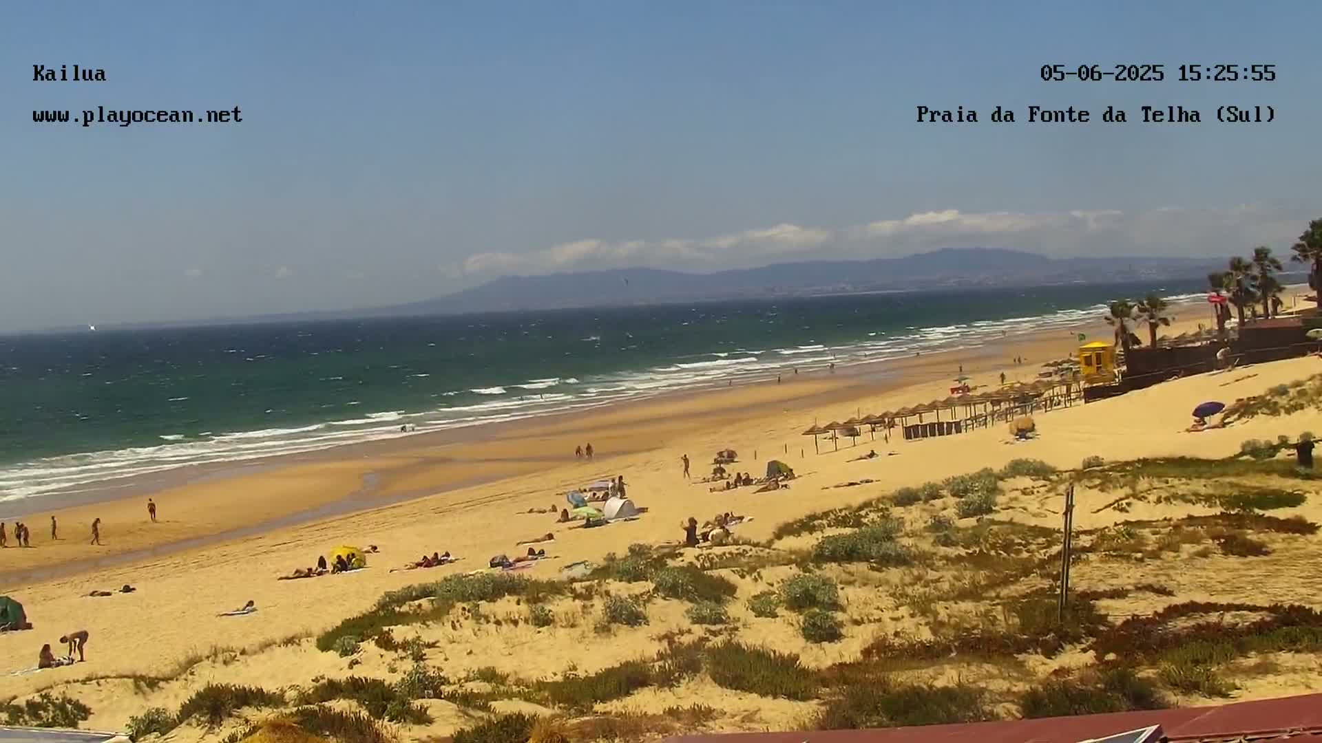 A sandy beach with scattered people and umbrellas under a mostly sunny sky, with a moderate surf rolling in from a teal ocean.