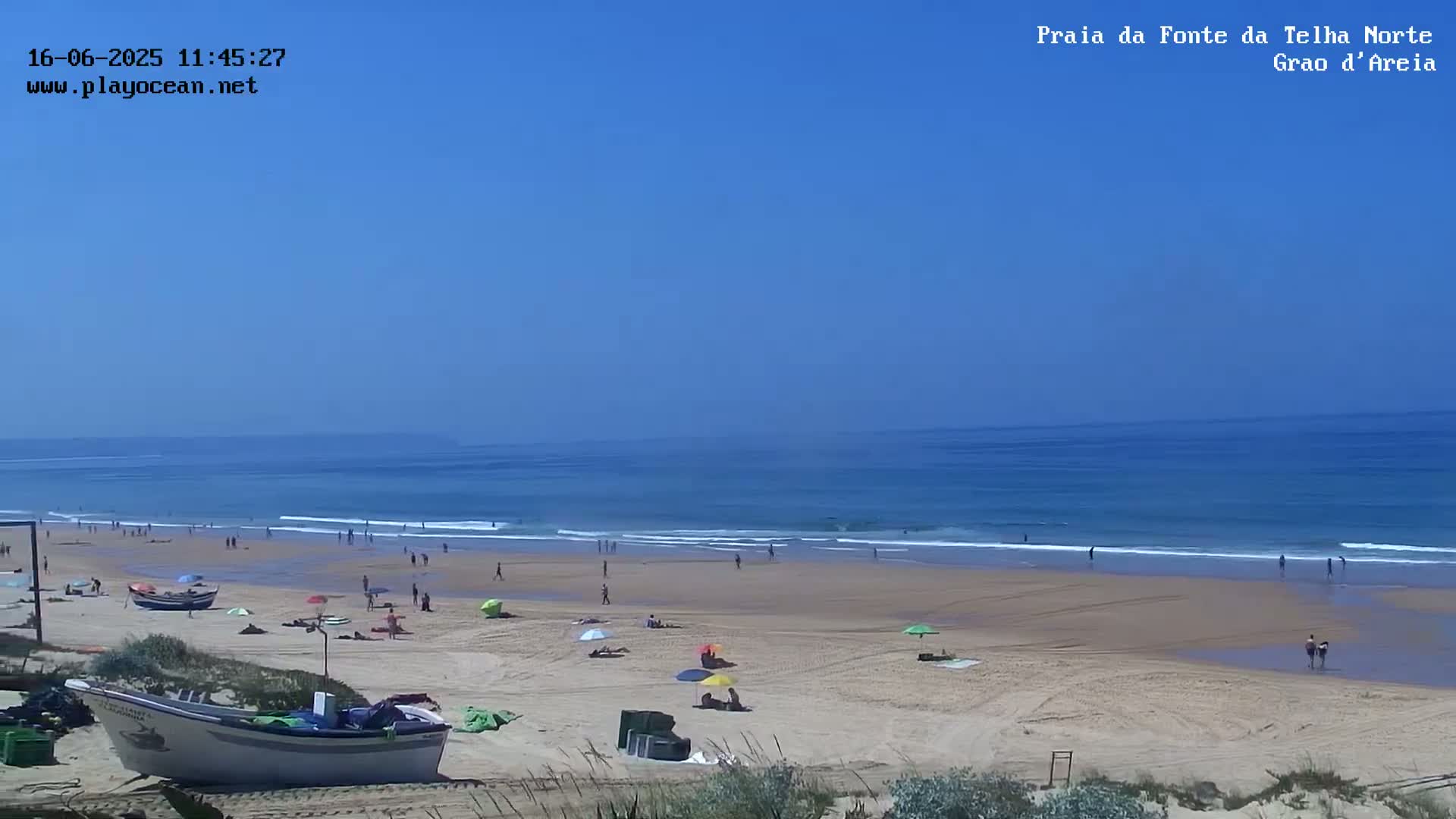 A sandy beach with scattered sunbathers and small waves under a clear blue sky is visible from a dune overlooking a small fishing boat.