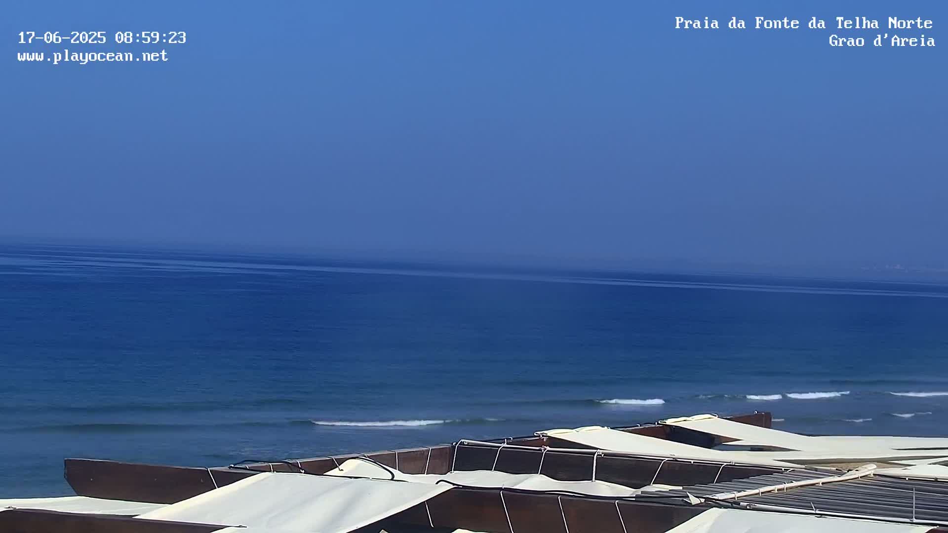 Under a clear blue sky, several dark wooden beach loungers with white canopies are situated overlooking a calm, deep blue ocean with small waves.