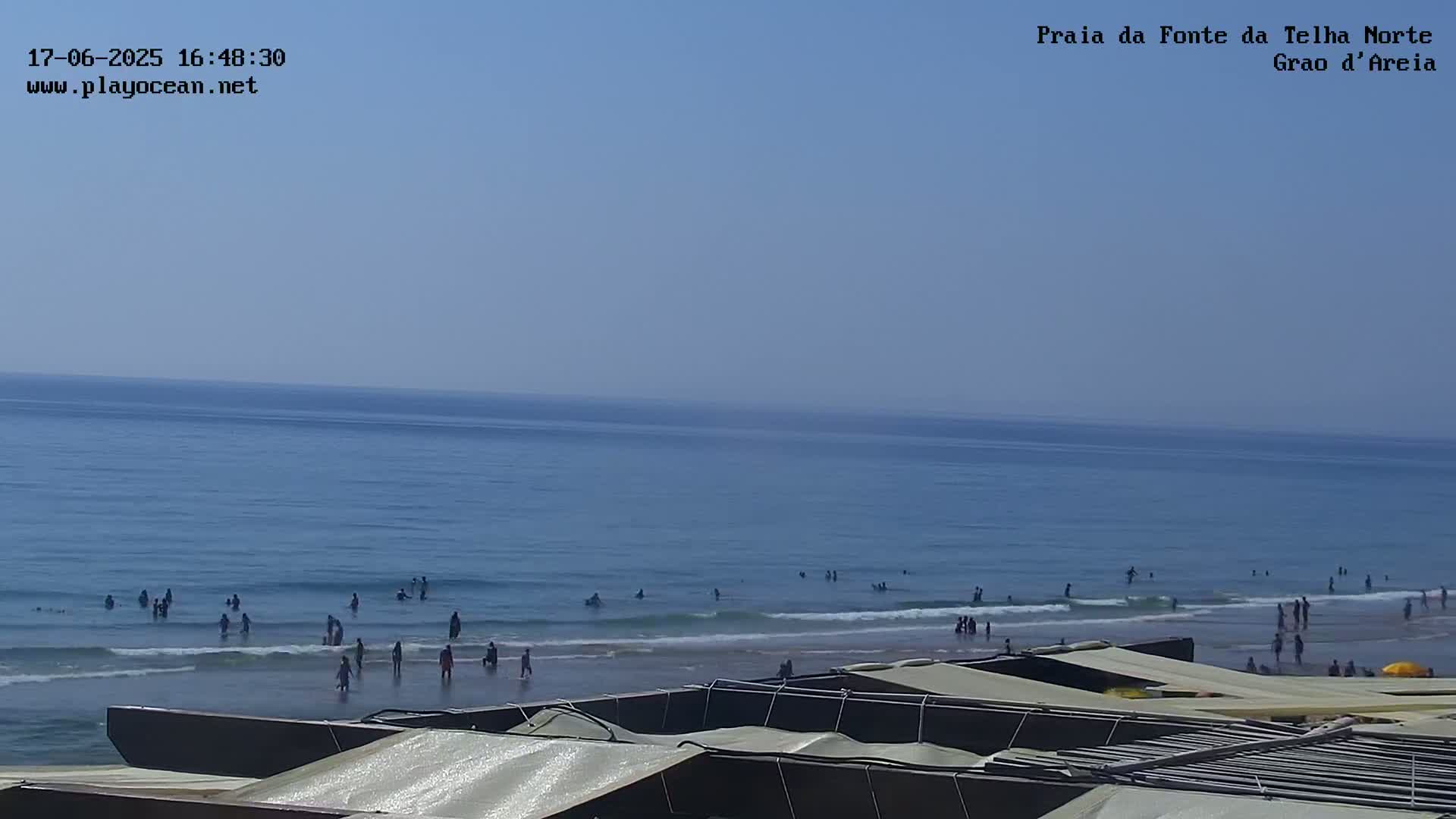 A mostly sunny day at a beach shows people swimming and wading in the ocean, with beach umbrellas and other structures in the foreground.