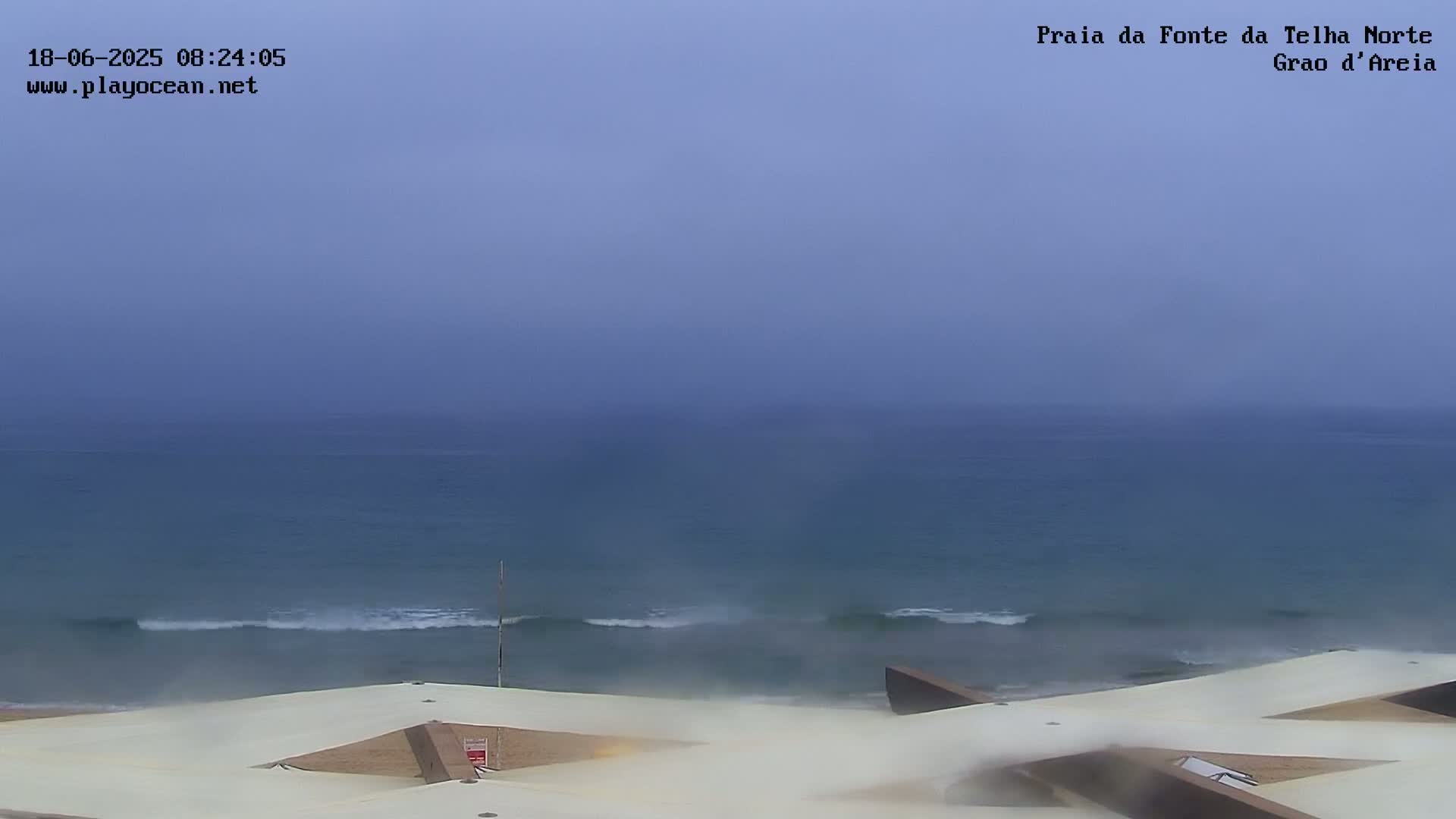 A hazy, overcast day reveals a calm ocean with small waves gently rolling onto a sandy beach, viewed from an elevated perspective of a white, geometric structure.