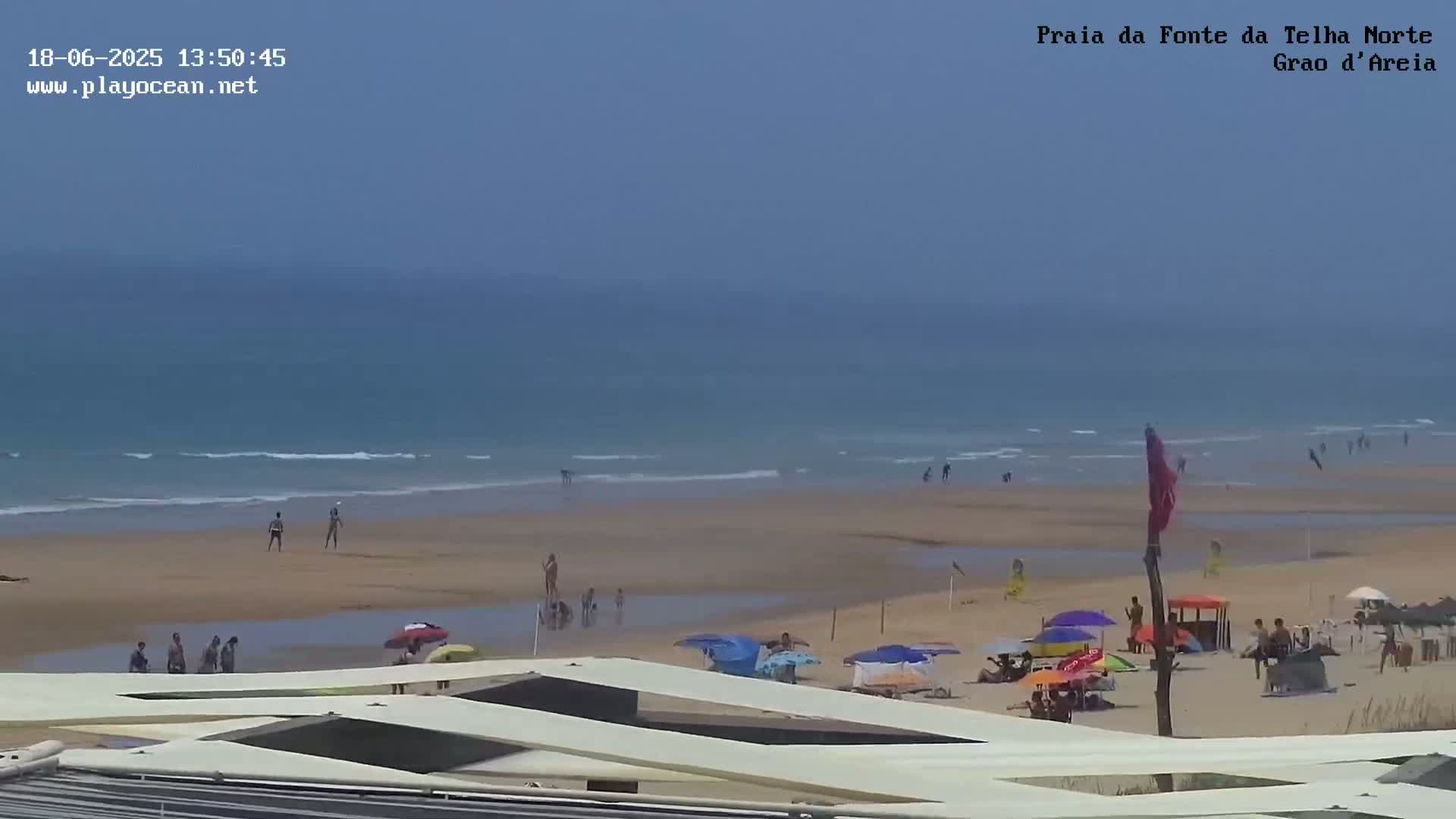 A sandy beach with people swimming and sunbathing under a hazy sky is seen from a high vantage point.