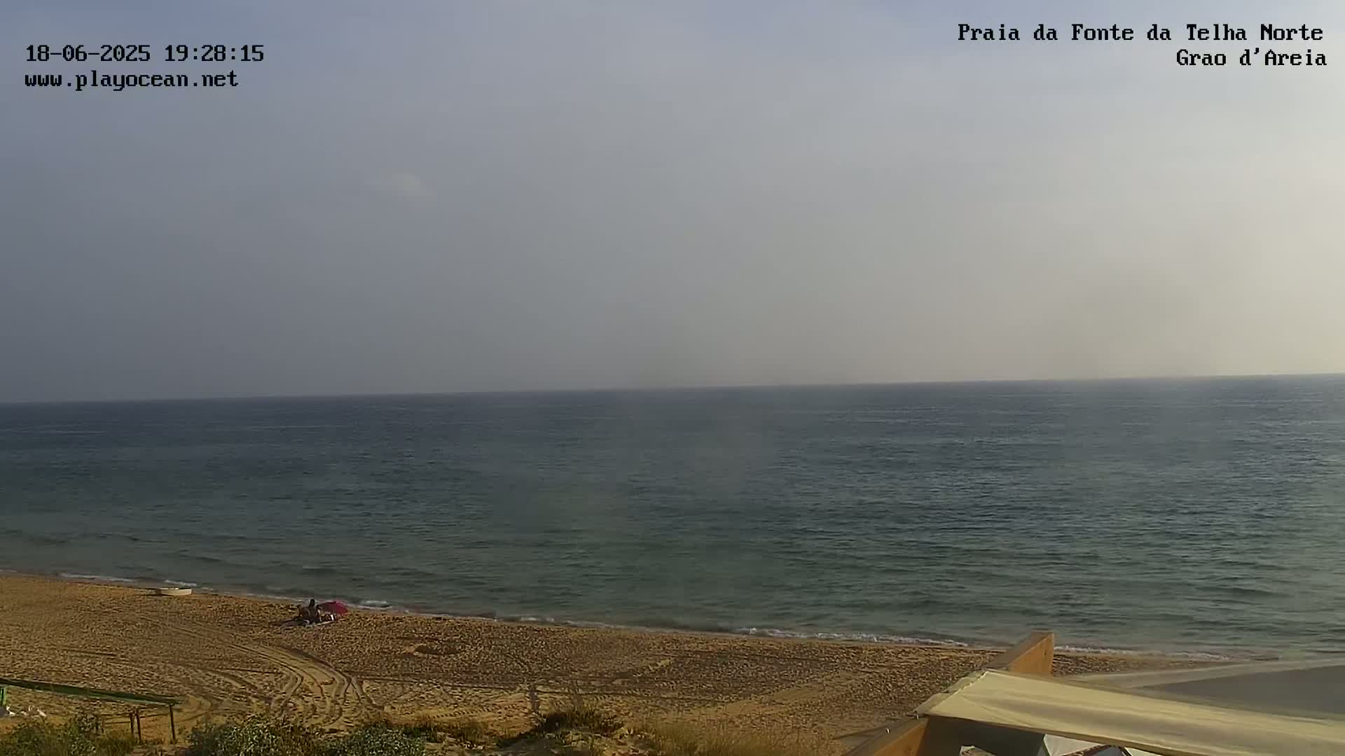 A mostly clear, hazy day at a sandy beach with calm ocean water and a few people visible near the shore.