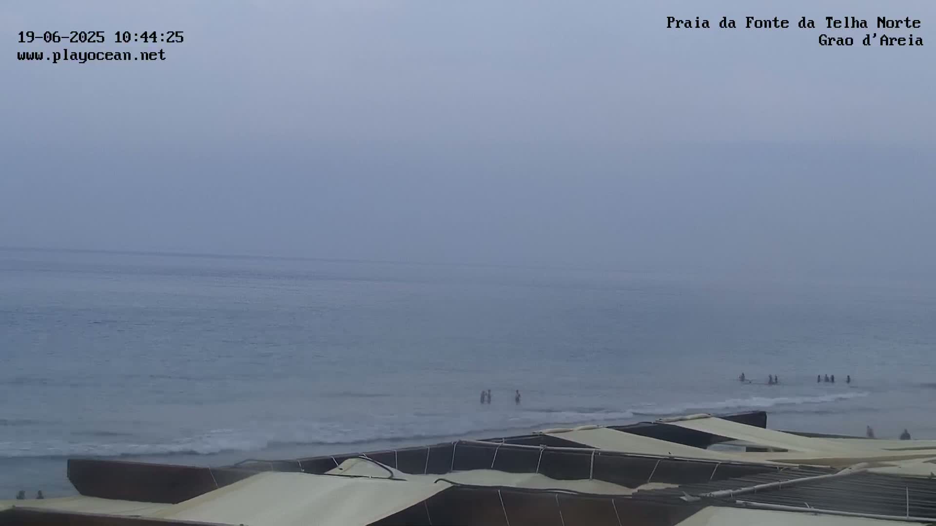 A hazy, overcast day at a beach shows calm ocean waves and several people wading in the water near the shore, with beach umbrellas in the foreground.