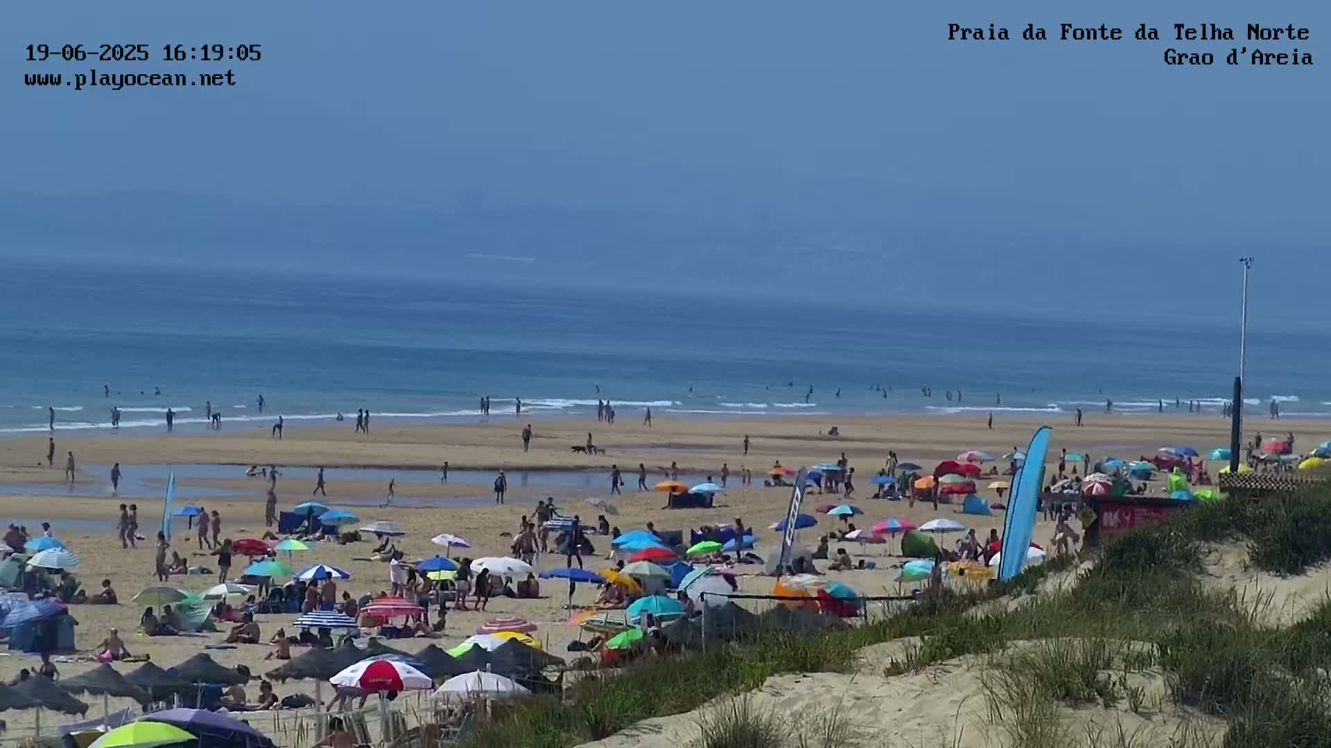 A crowded beach with many colorful umbrellas and people under a clear, sunny sky, with calm ocean waves in the background.