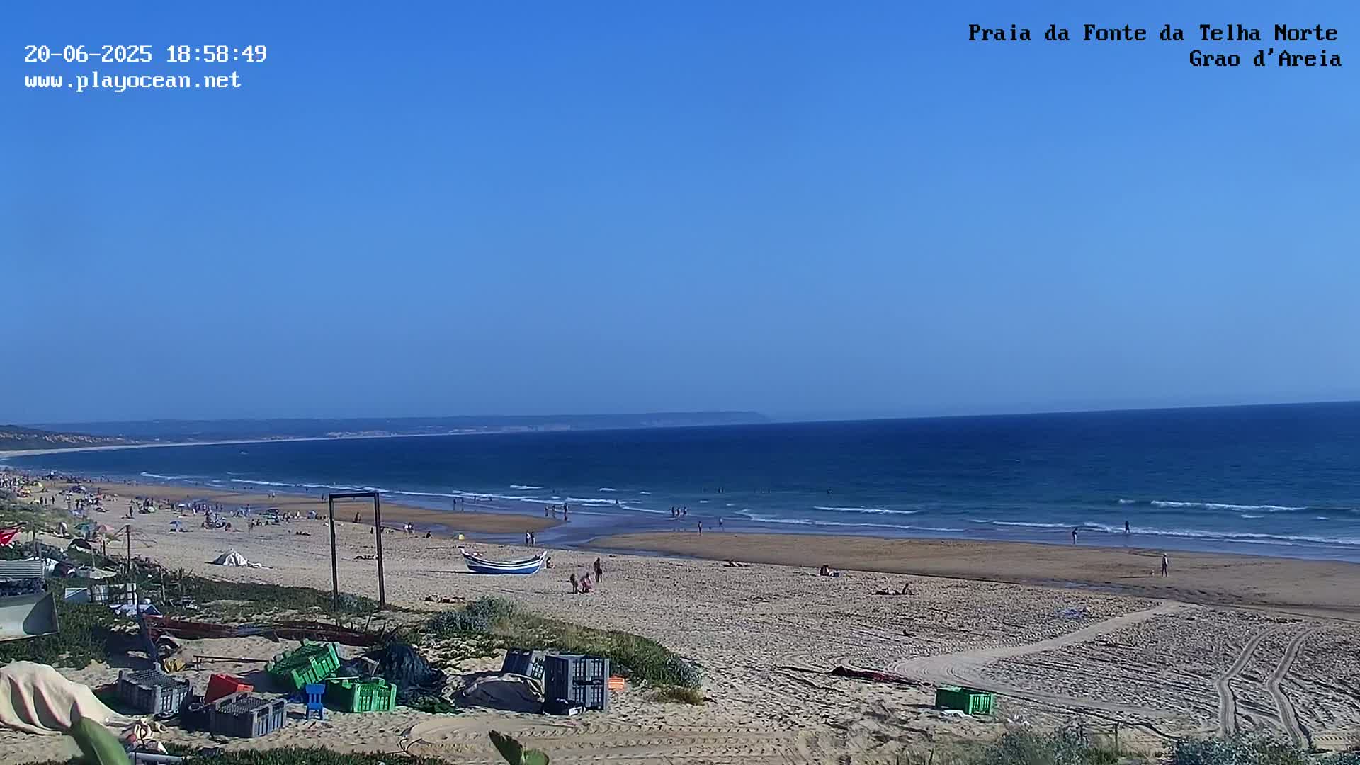 A sandy beach with scattered people and a calm ocean under a clear blue sky is seen from a slightly elevated vantage point.