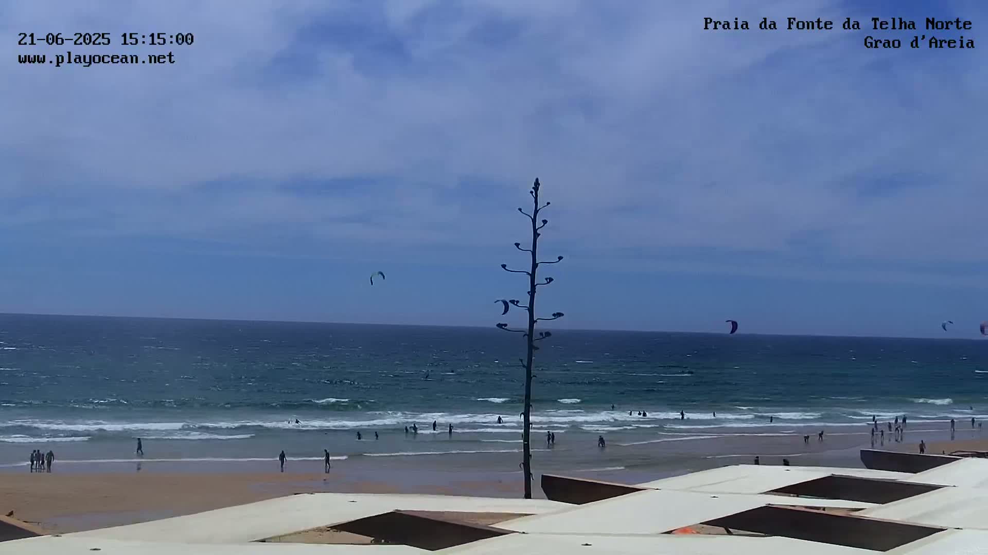 A mostly sunny day at a beach shows a few people swimming and walking on the sand, with several kitesurfers in the ocean, all viewed from atop a structure with white coverings.