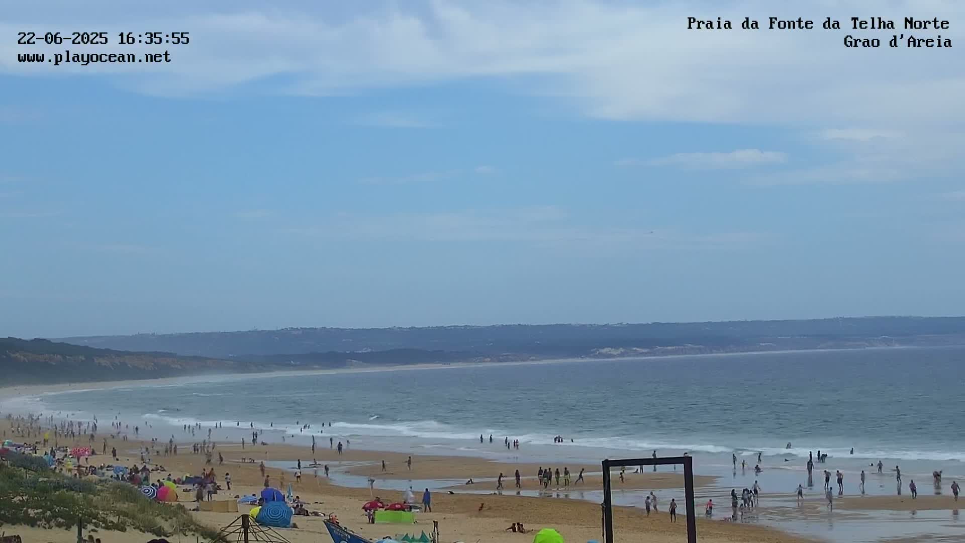 A sandy beach with many people on it and in the water stretches along a calm ocean under a mostly clear, light blue sky.