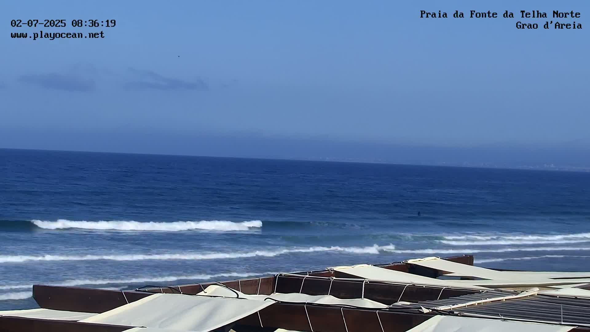 Under a clear blue sky, several dark-brown, covered beach loungers are situated overlooking a calm ocean with small waves breaking gently near the shore.