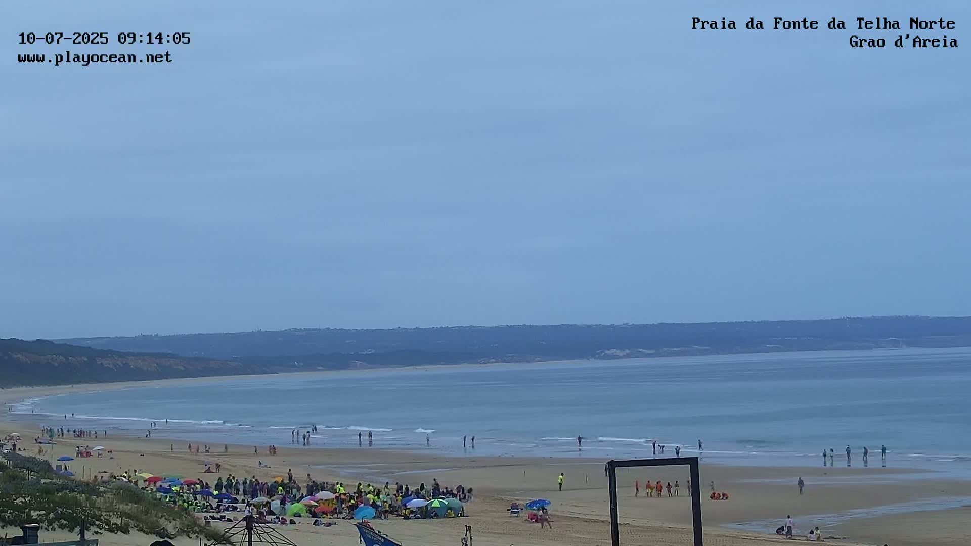 A sandy beach with many people enjoying the sunny day, under a clear, light-blue sky, next to a calm ocean with small waves.