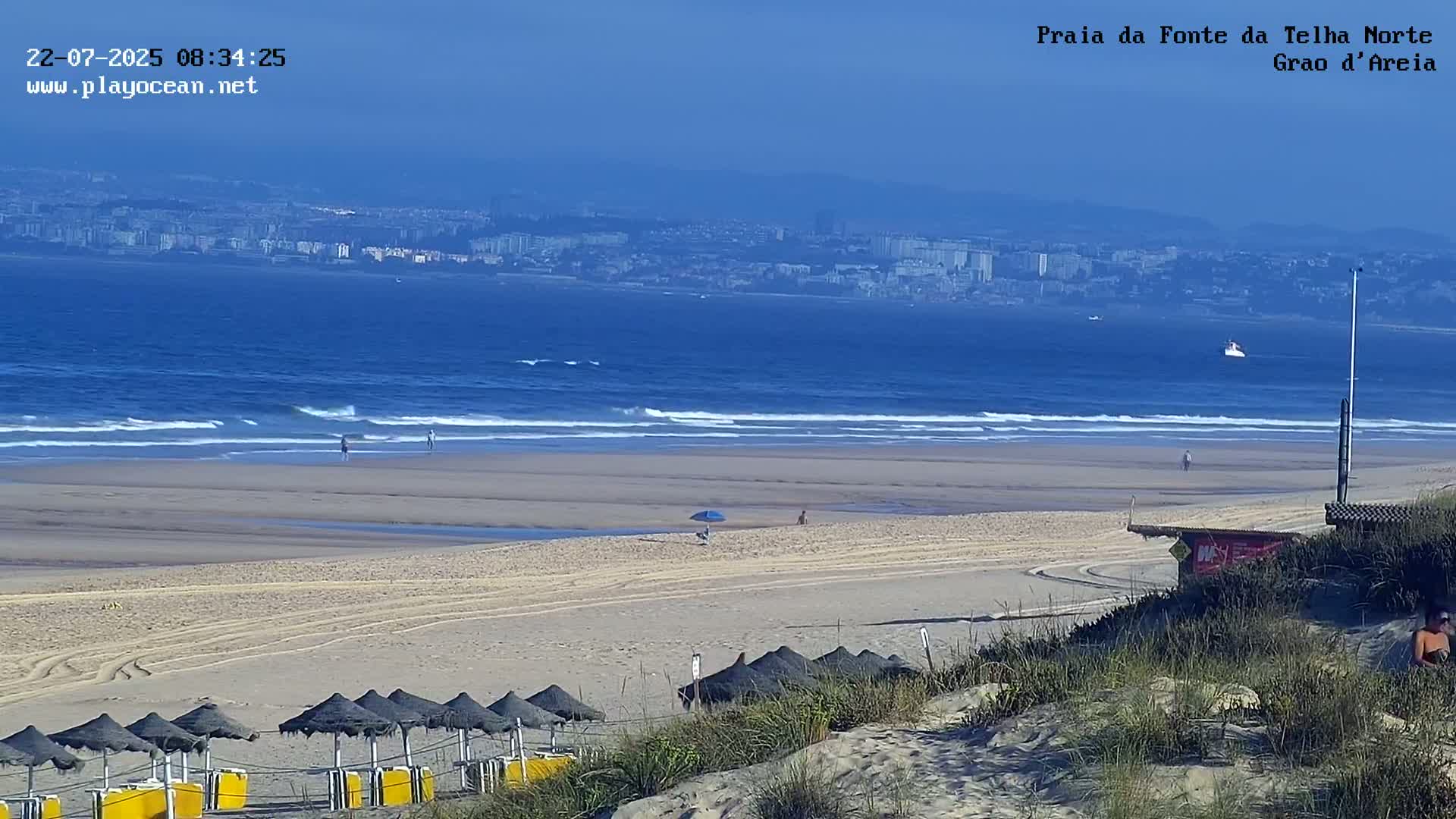 A sandy beach with small waves, several people, and rows of closed beach umbrellas stretches toward a distant city under a clear blue sky.