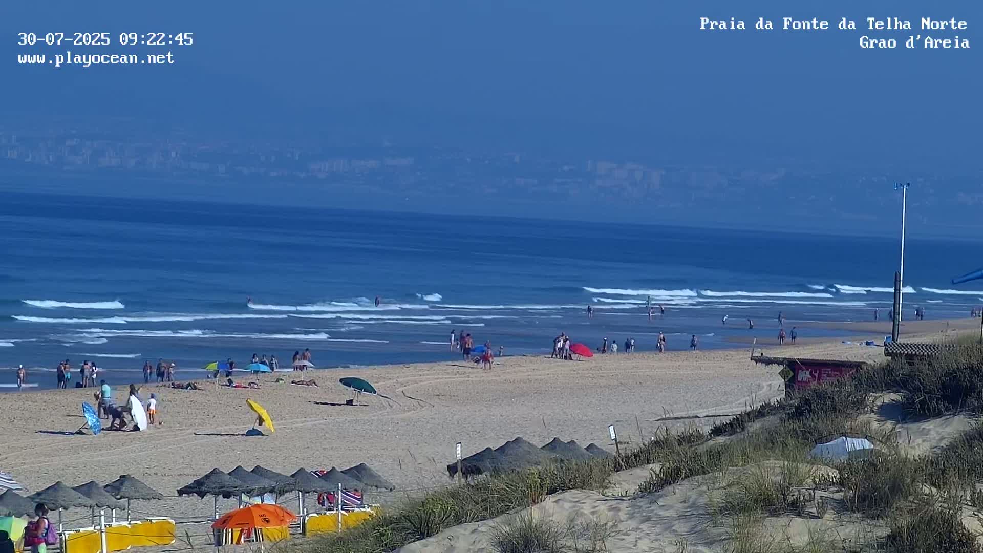 A sandy beach with small waves, numerous people, and beach umbrellas, overlooks a calm ocean under a clear blue sky.