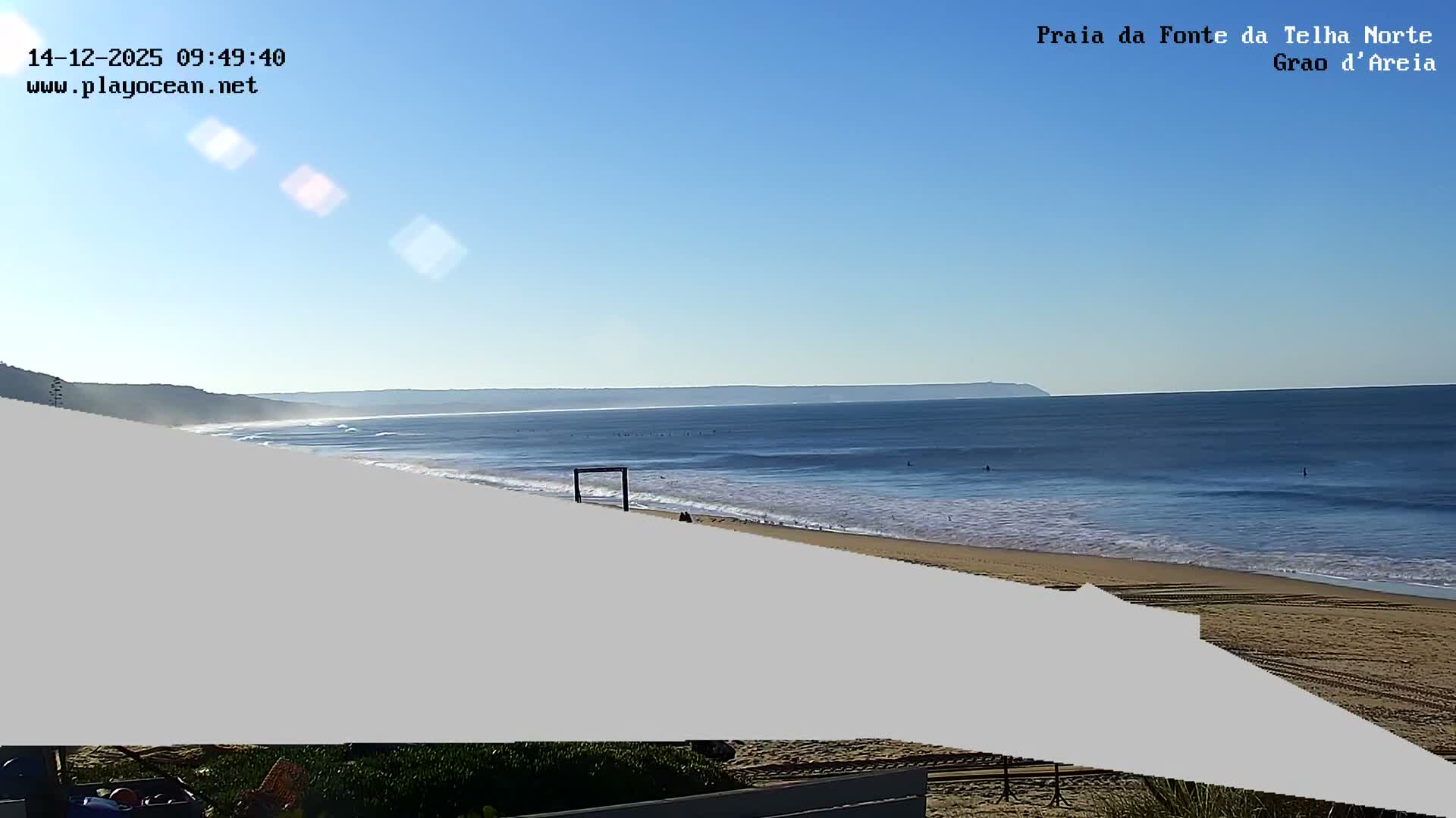 A sandy beach with small waves, numerous people, and beach umbrellas, overlooks a calm ocean under a clear blue sky.