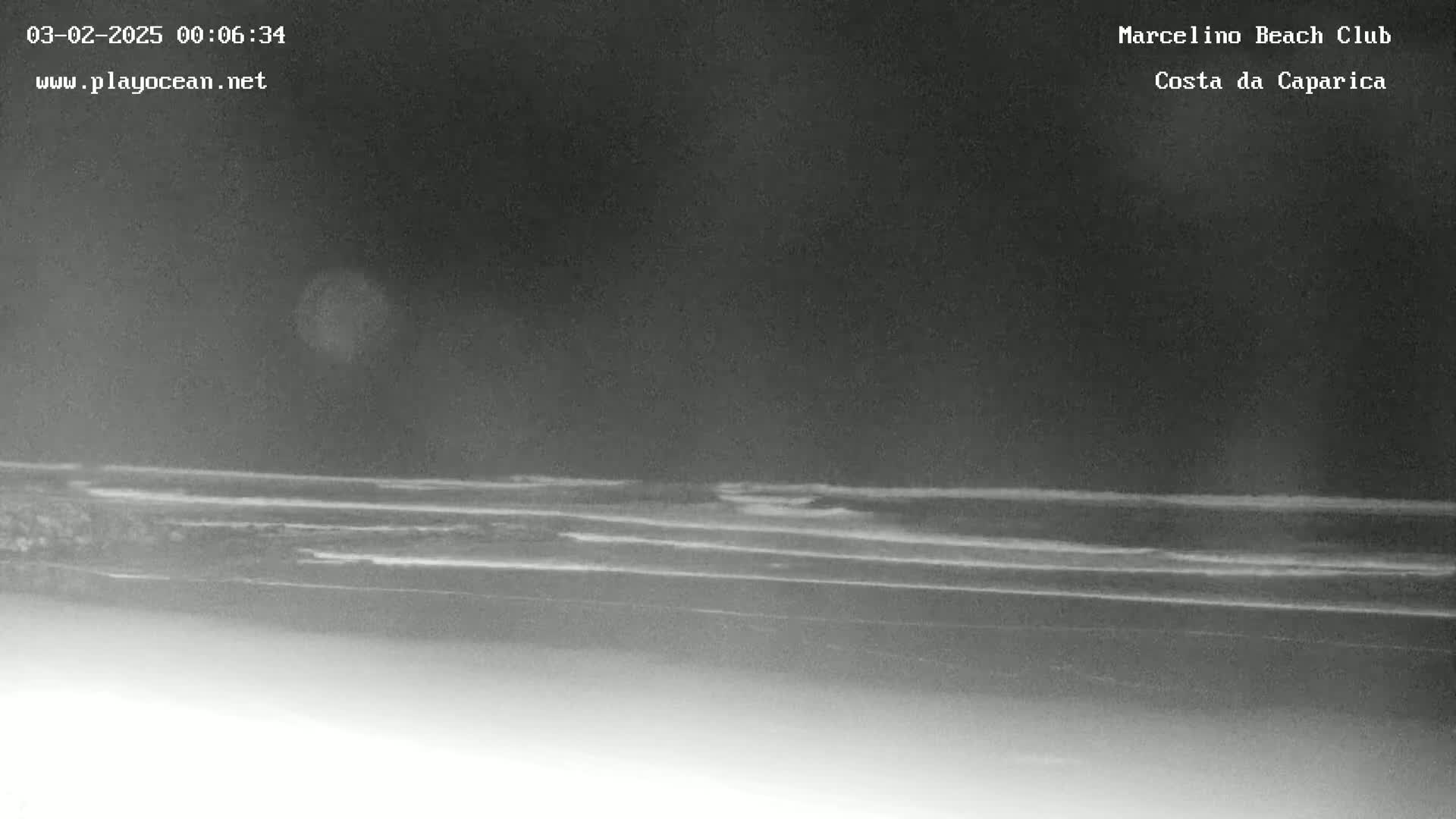 A nighttime, low-light grayscale image shows dark ocean waves lapping a beach under a mostly cloudy sky.