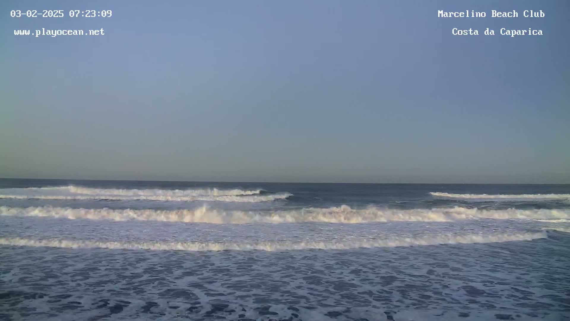Ocean waves are breaking on a shore under a clear, light-blue sky.