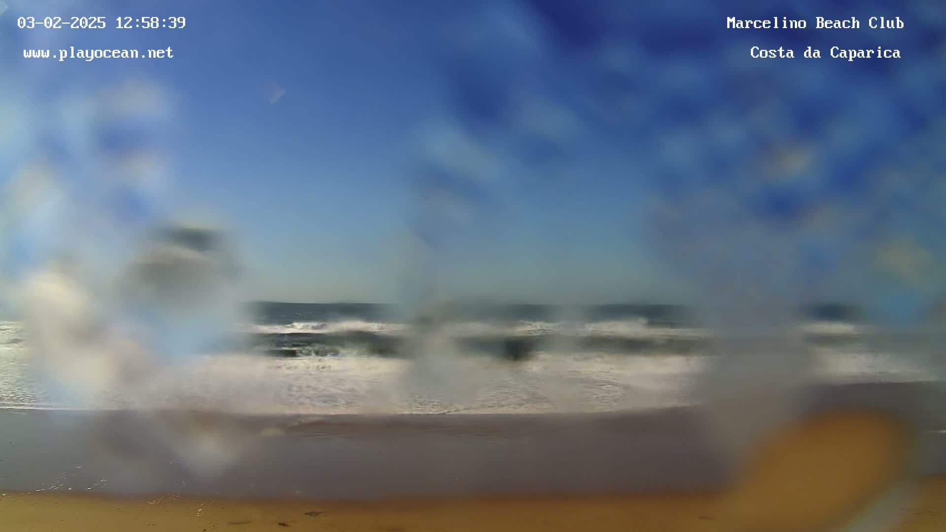 A blurry view of an ocean beach on a clear day, showing waves breaking on the shore and a sandy beach in the foreground.