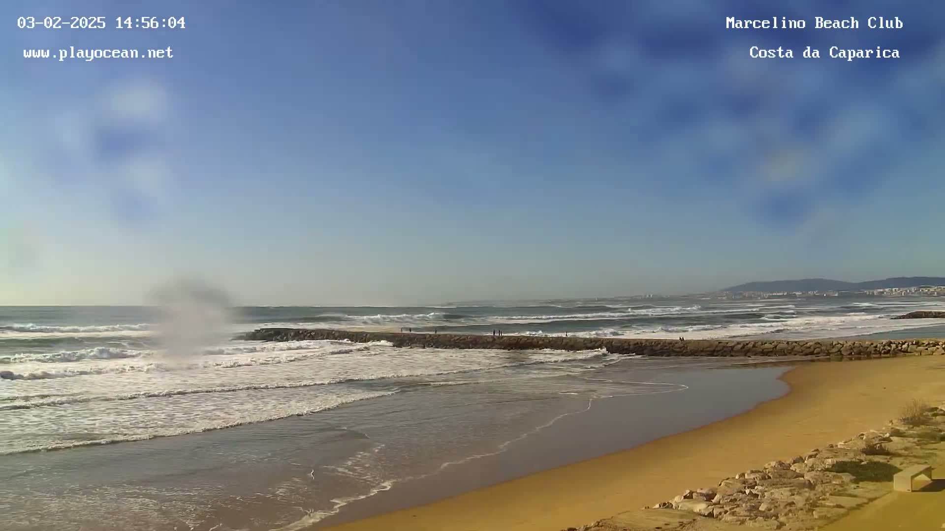 A sandy beach with a rock jetty extending into a wavy ocean under a clear blue sky.