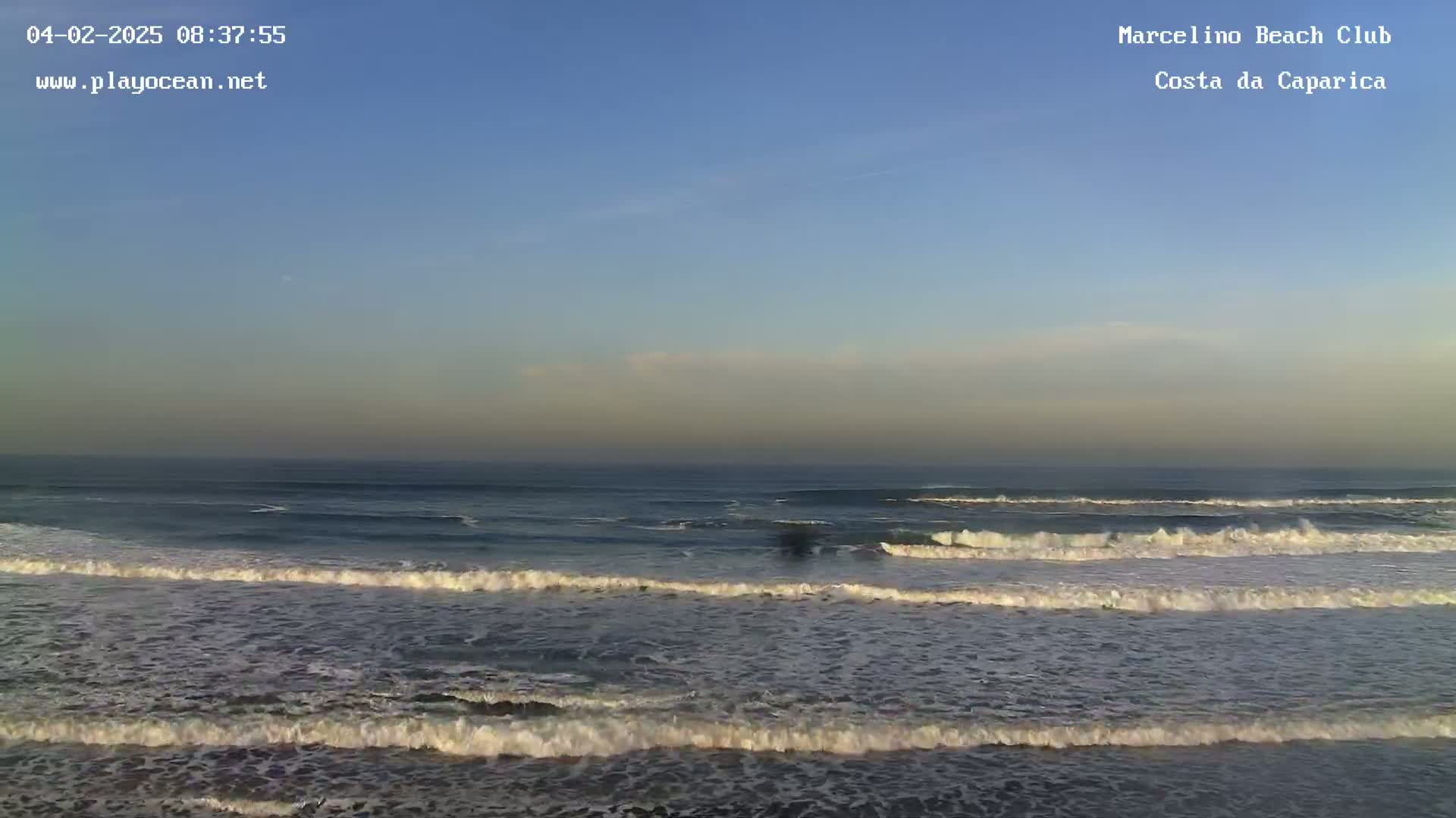 A wide shot of ocean waves rolling onto a beach under a clear, pale blue sky.