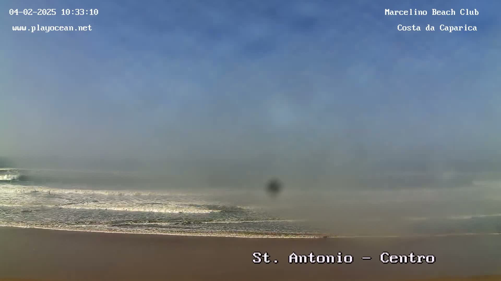 A hazy, mostly clear day shows a beach with waves gently rolling onto the shore.