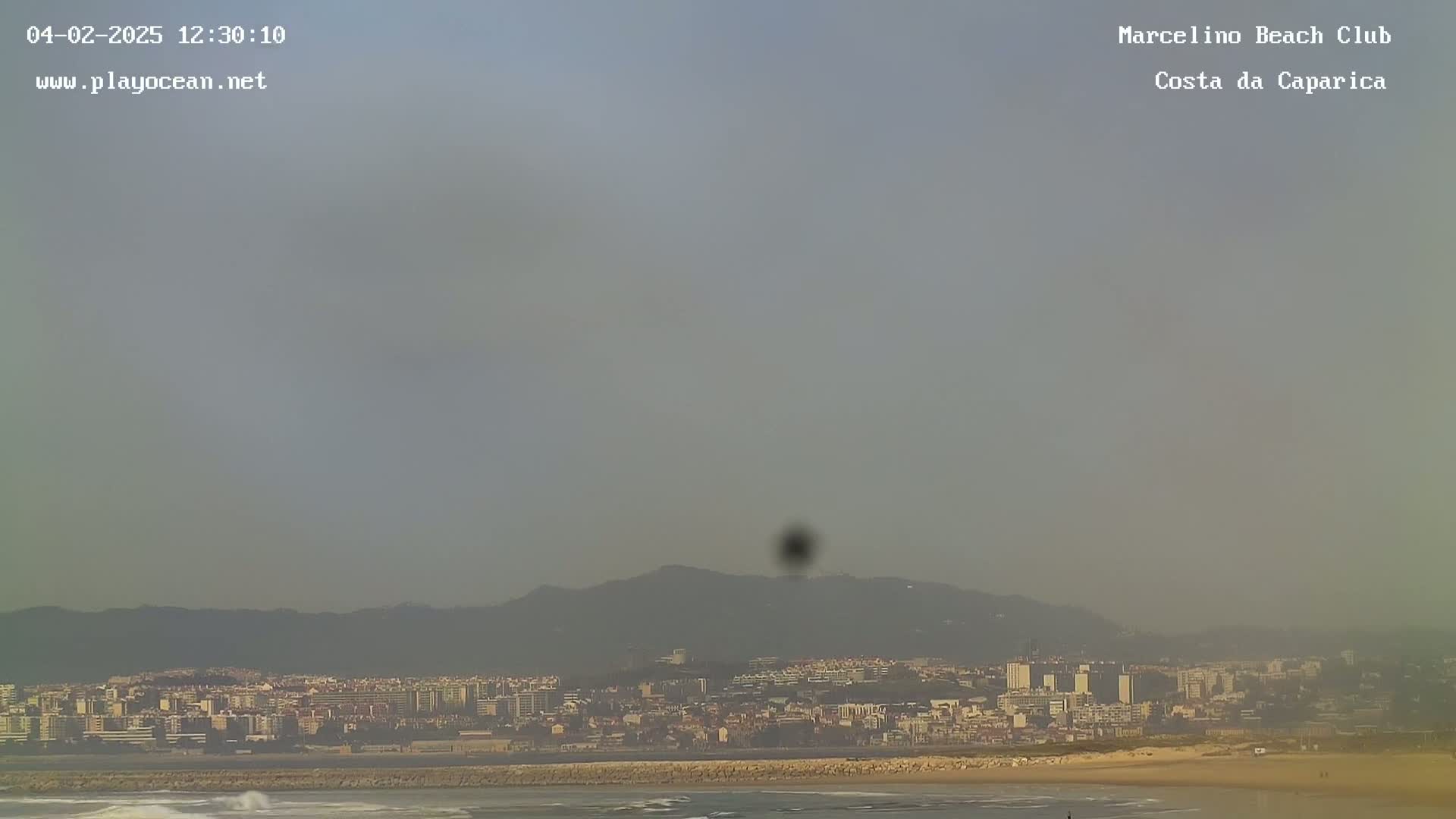 A hazy view shows a beach, ocean, and a city nestled against distant mountains.