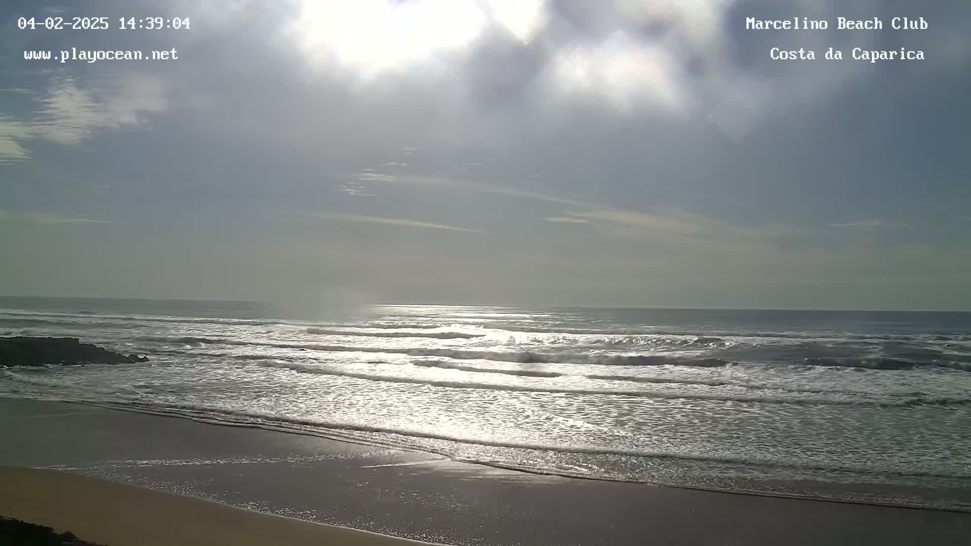 A mostly cloudy sky brightens over a sandy beach where waves roll onto the shore.