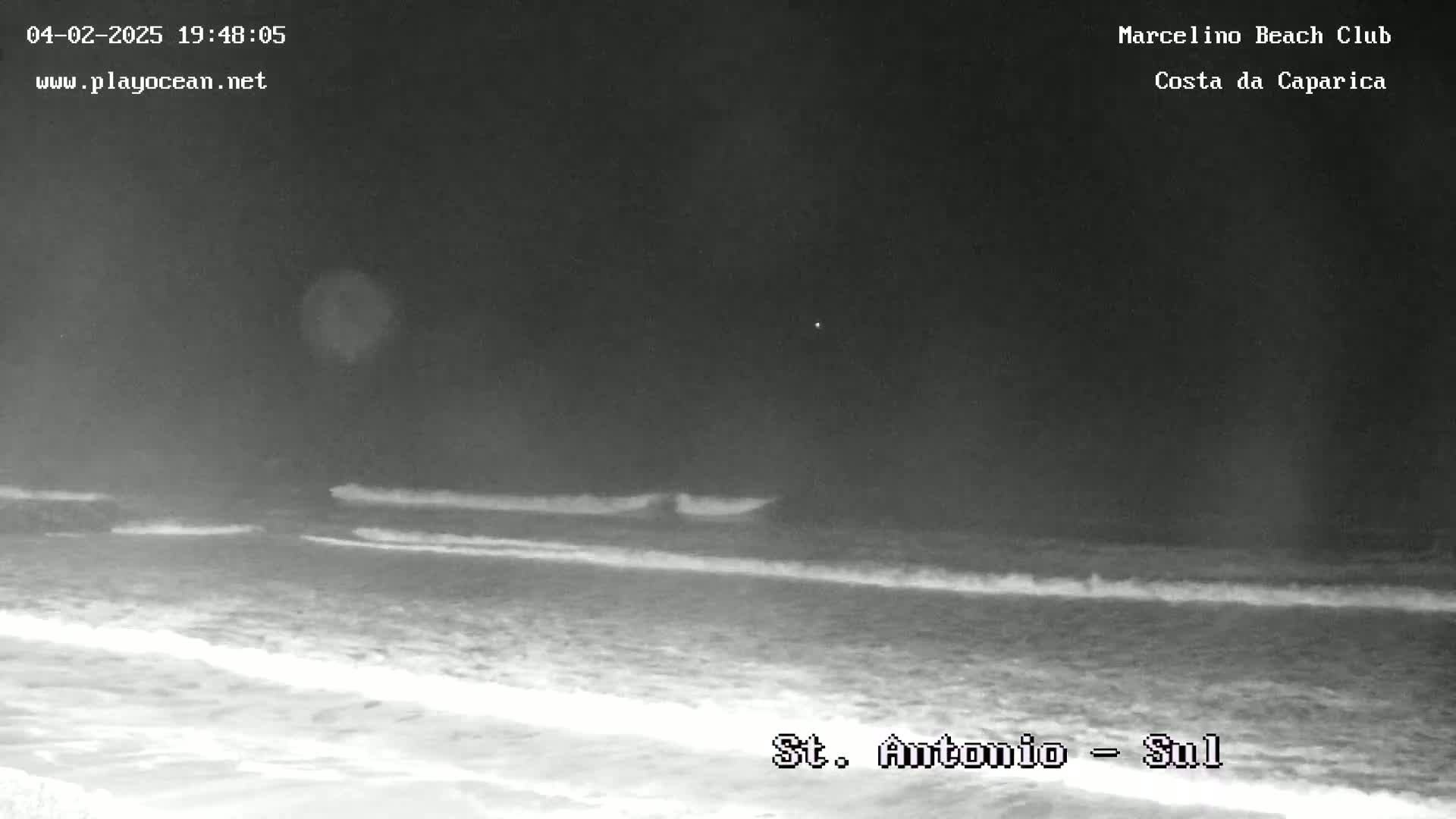 A nighttime, low-light beach scene shows gentle waves lapping the shore under a dark sky.