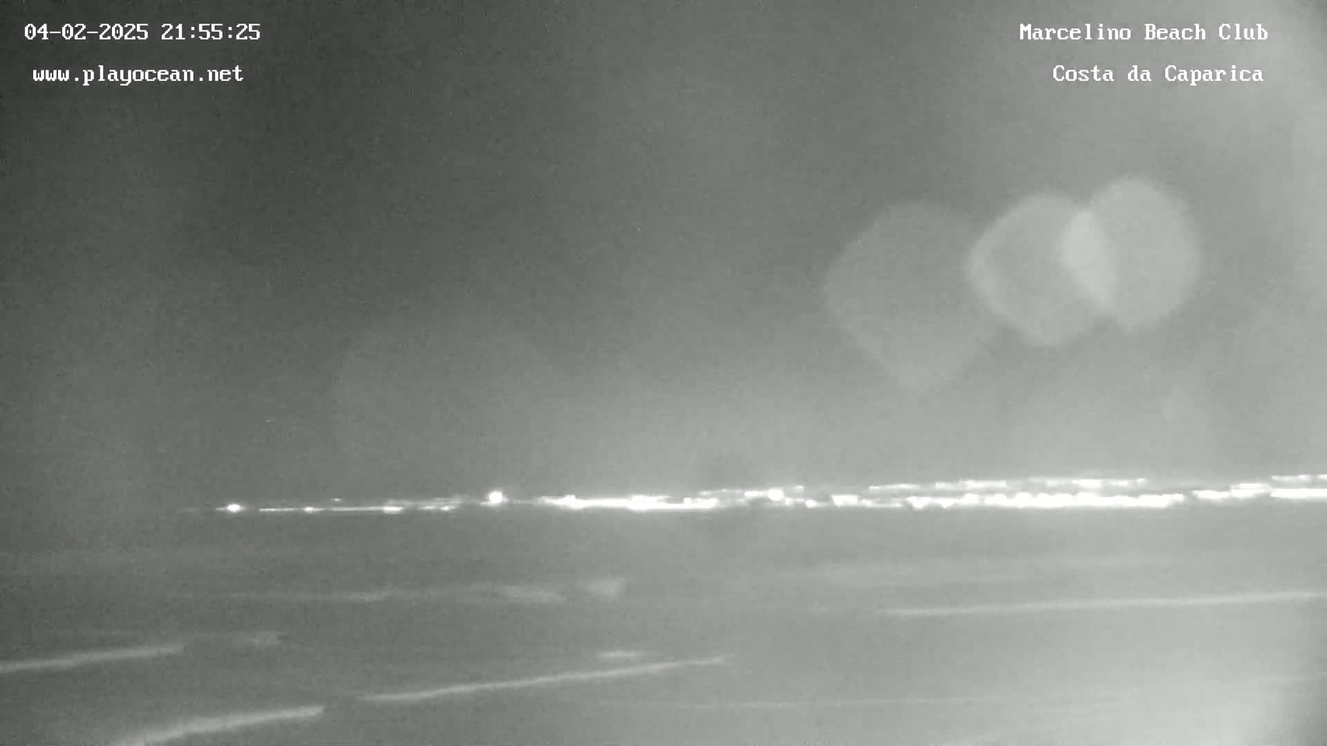 A nighttime long shot of a coastline shows a faint line of city lights on the horizon under a dark, overcast sky.