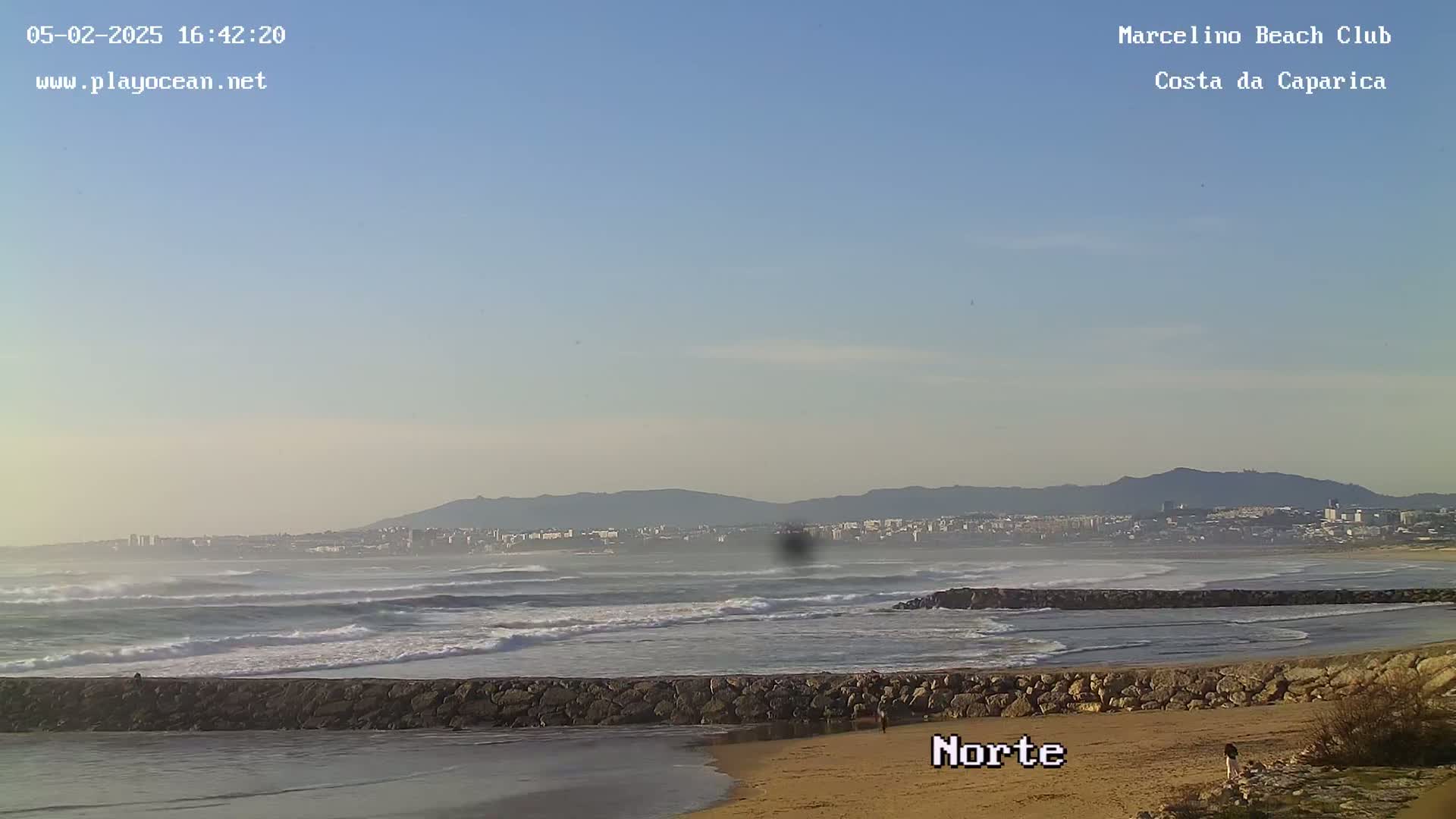 A sandy beach with a rocky barrier separating a calm inlet from a moderately wavy ocean, under a clear, light blue sky, with a distant city and mountains visible on the horizon.