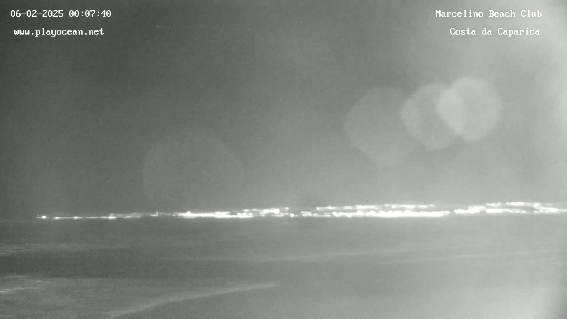 A nighttime, low-light view of a dark beach and ocean with a distant line of lights on the horizon.