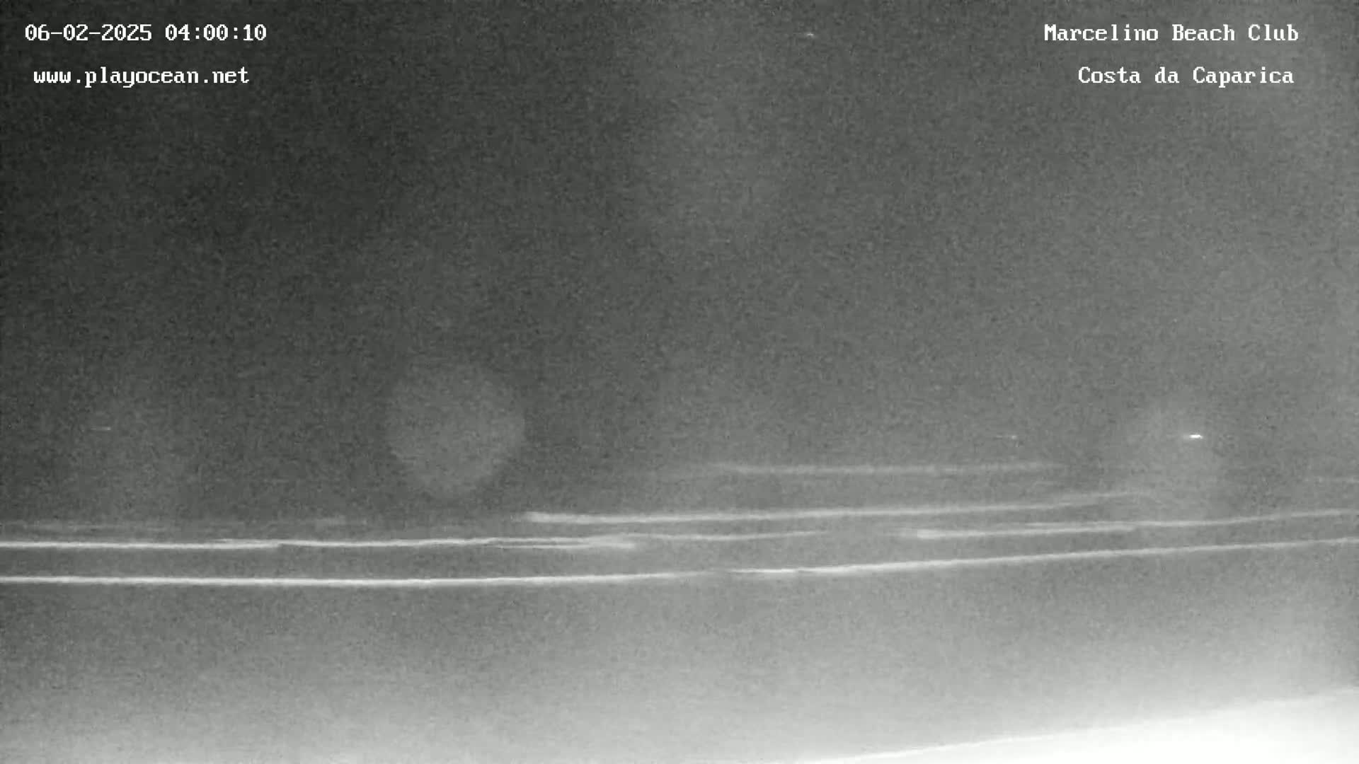 A nighttime, low-light image shows a dark beach with faintly visible waves and a dark, hazy sky.