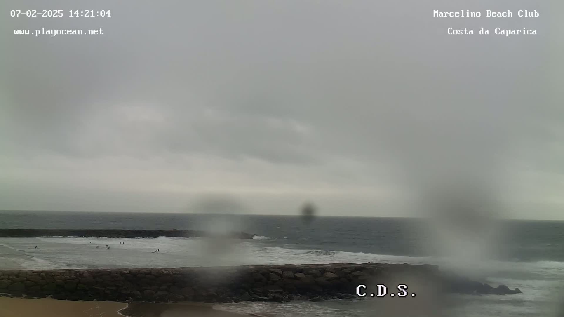 A cloudy, overcast day at a beach shows a calm ocean with several people in the water near a stone wall.
