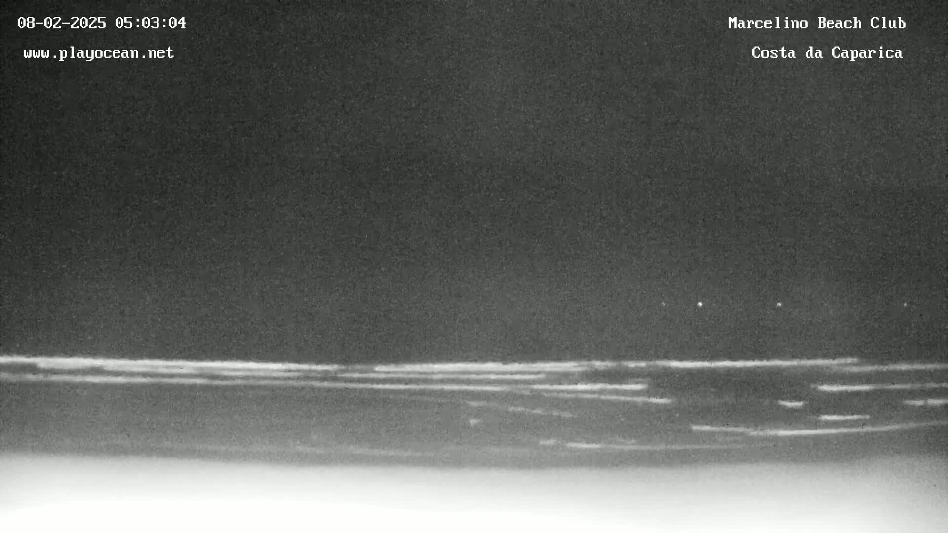 A dark, nighttime beach scene shows a dark ocean with faint, horizontal lines likely representing waves under a mostly clear sky.