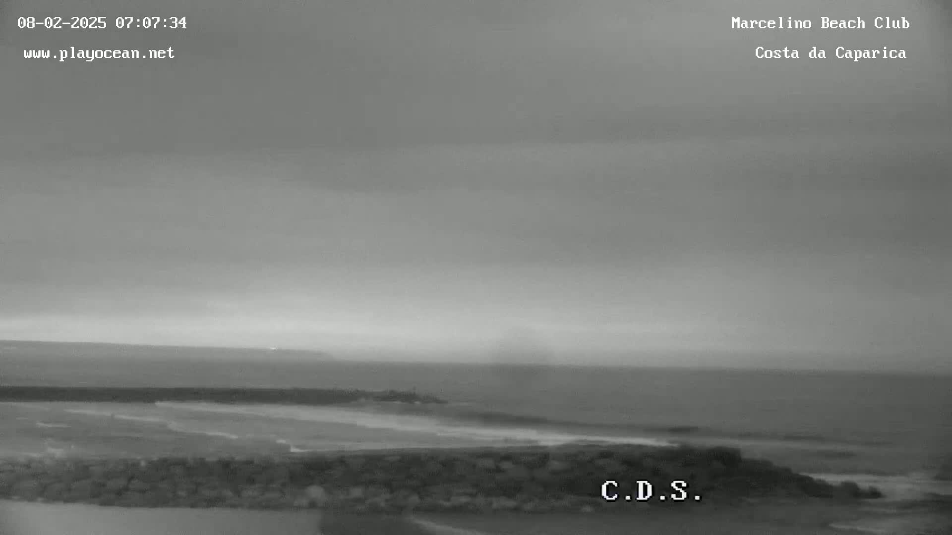 A mostly overcast sky hangs over a calm ocean meeting a sandy beach, partially obscured by a low rock wall.
