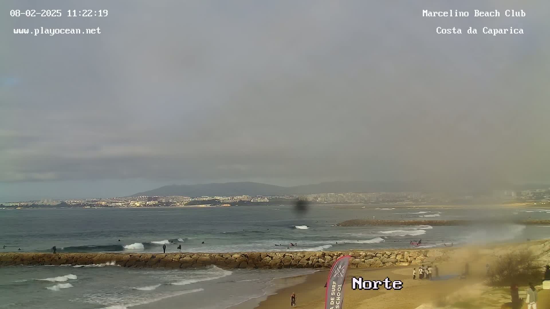 A mostly overcast day shows surfers in the ocean, waves breaking near a stone wall, and a sandy beach with a few people on it, all with a distant cityscape visible.