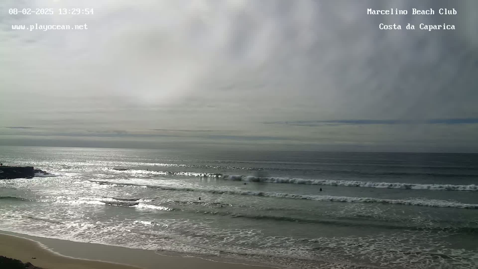 A mostly cloudy day shows a wide expanse of ocean with several surfers visible in the waves, along with a sandy beach in the foreground.