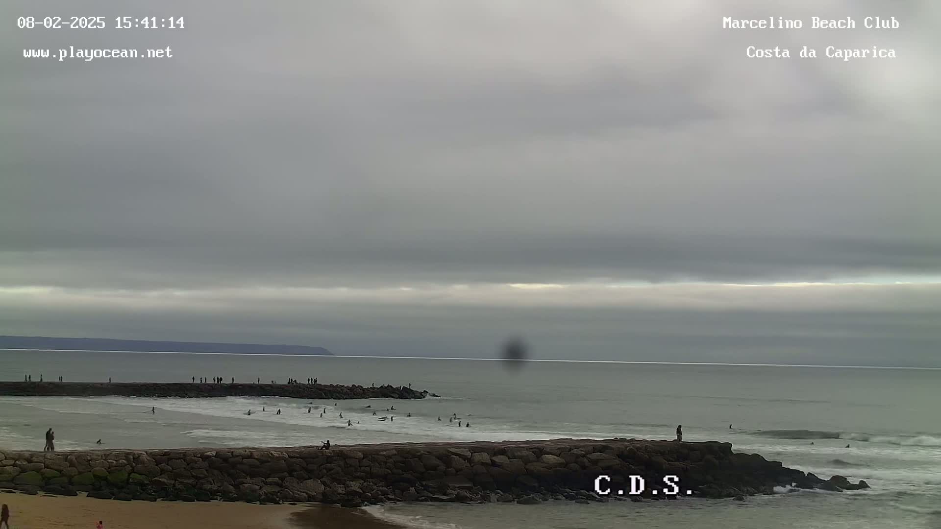 A mostly cloudy day at a beach shows a rocky jetty with people on it and in the water, and a calm ocean with a distant landmass.