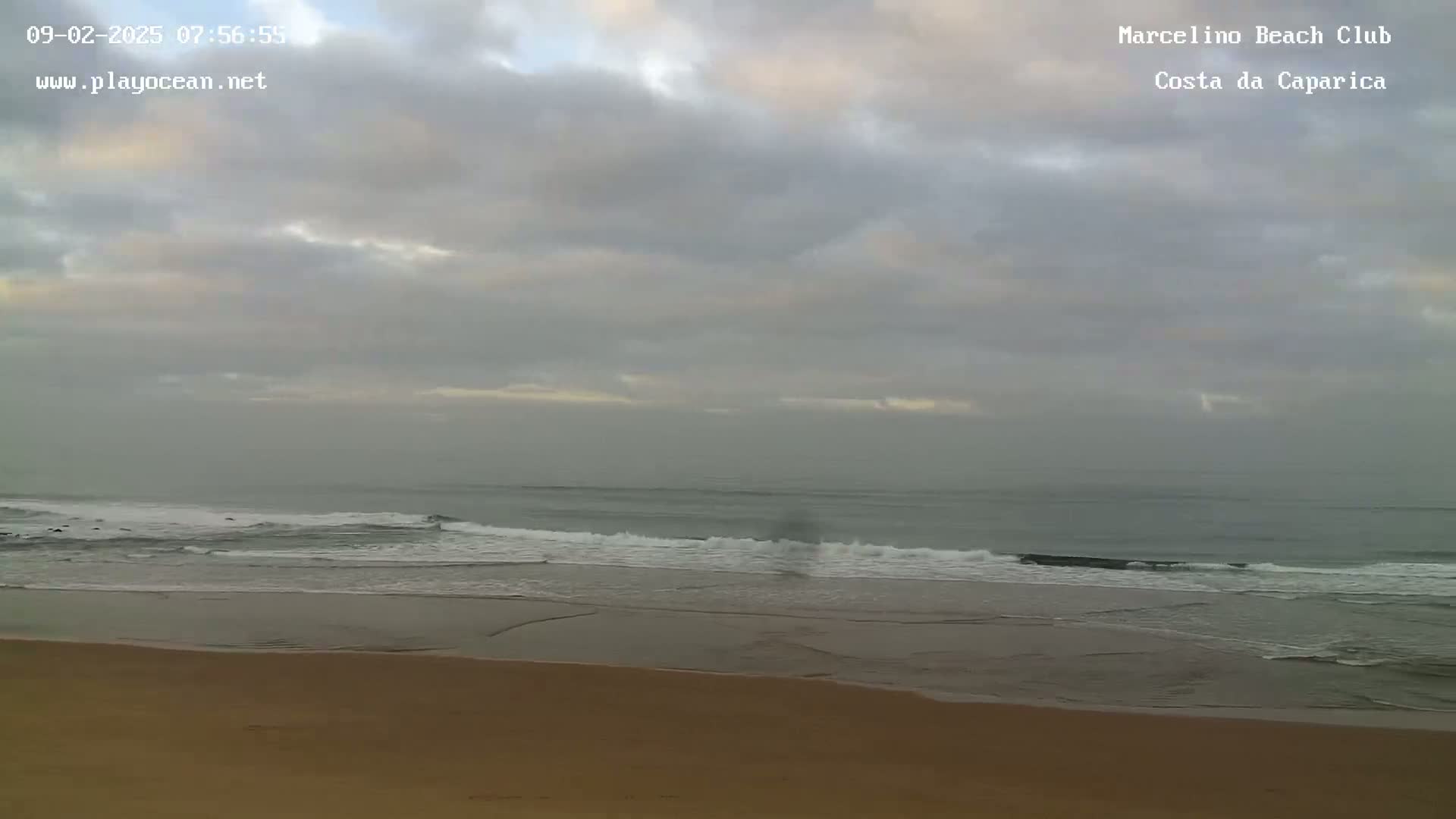 A sandy beach meets a calm ocean under a cloudy, overcast sky.