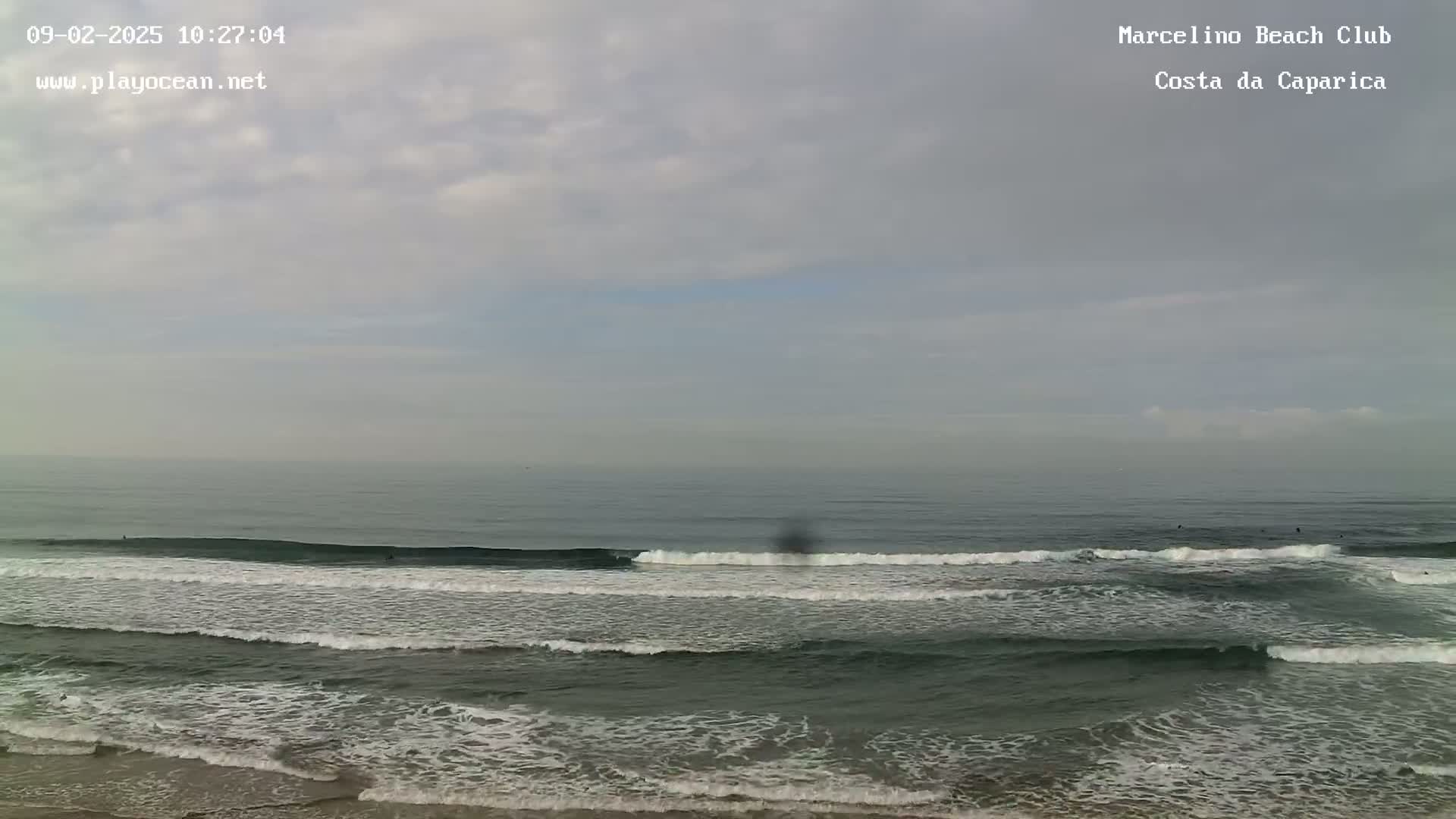 A mostly cloudy day shows an ocean with several surfers riding waves that are breaking near the shore.