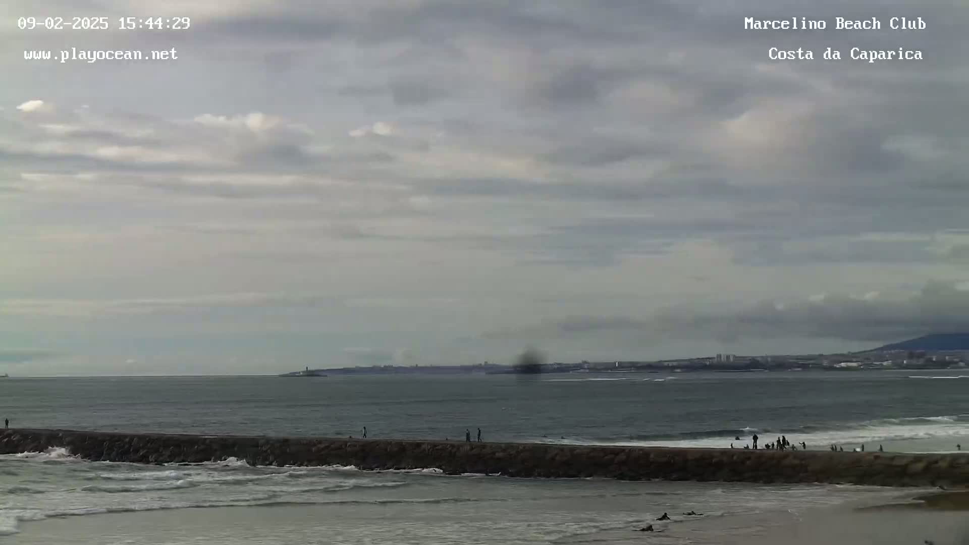 A long rock jetty separates a beach with several people from a gray ocean under a cloudy sky.