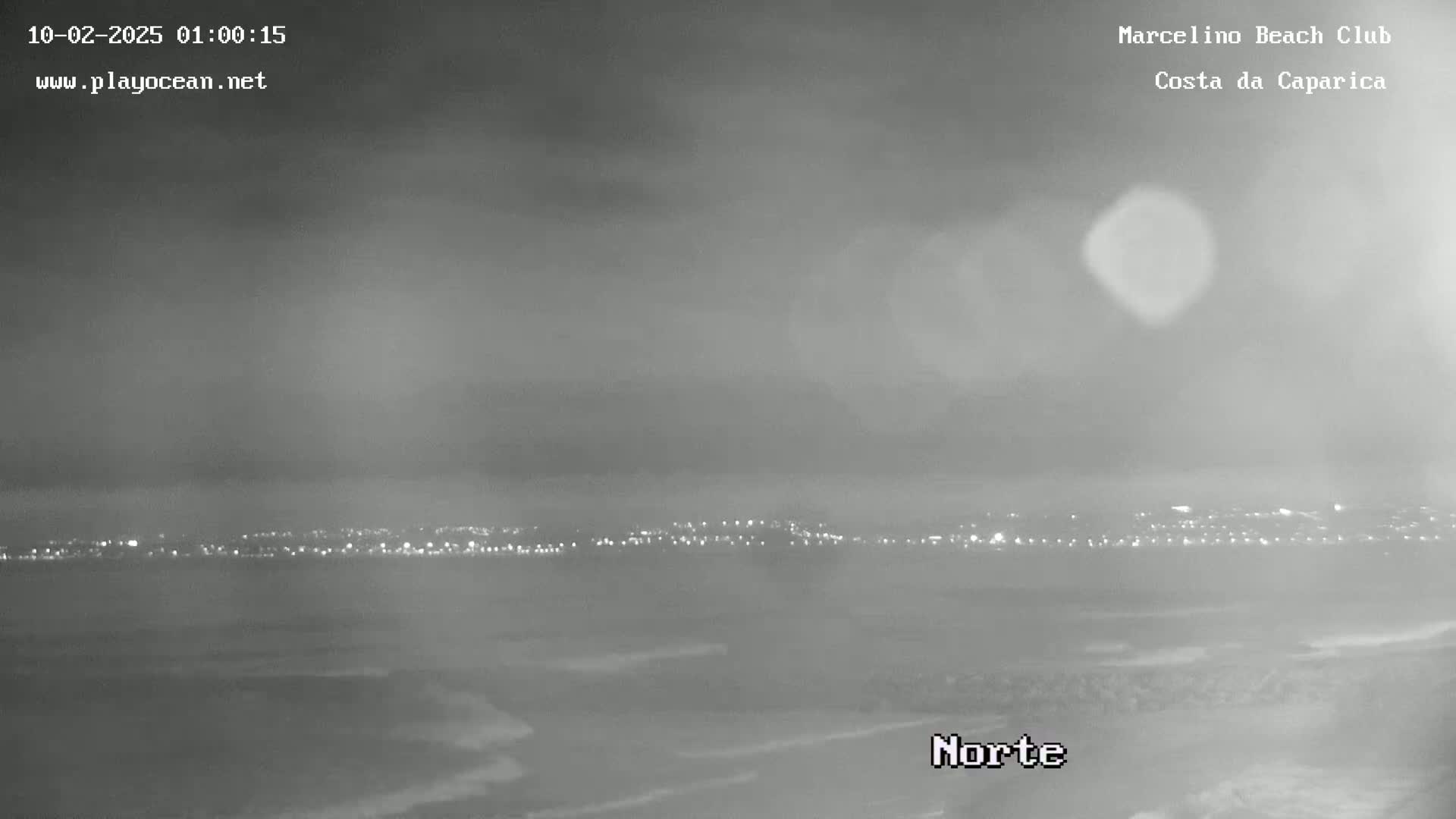 A nighttime, hazy view of a coastline shows a city's lights in the distance and dark water with faint waves in the foreground.