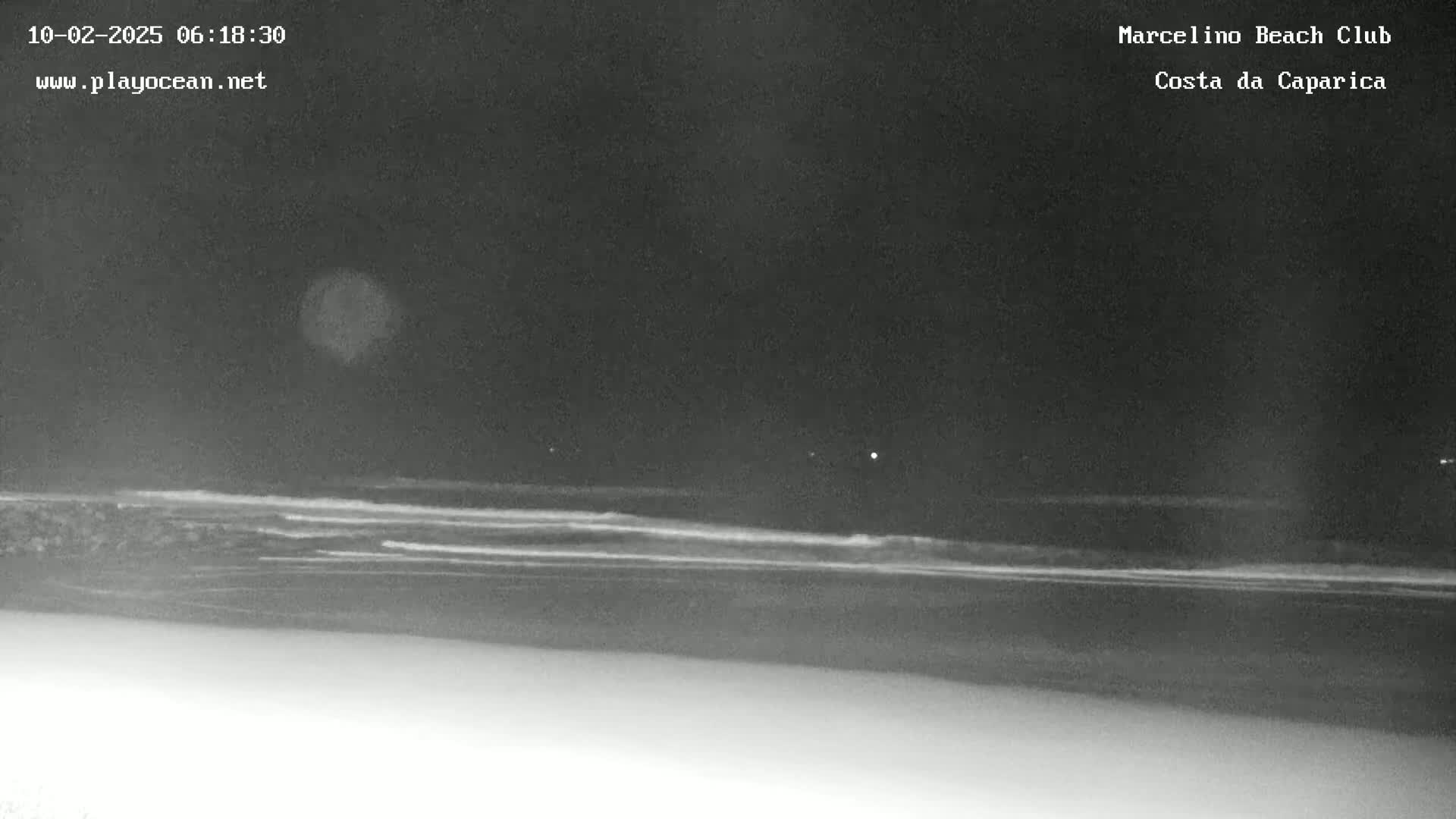 A nighttime, low-light image shows dark ocean waves lapping a sandy beach under a mostly dark sky.