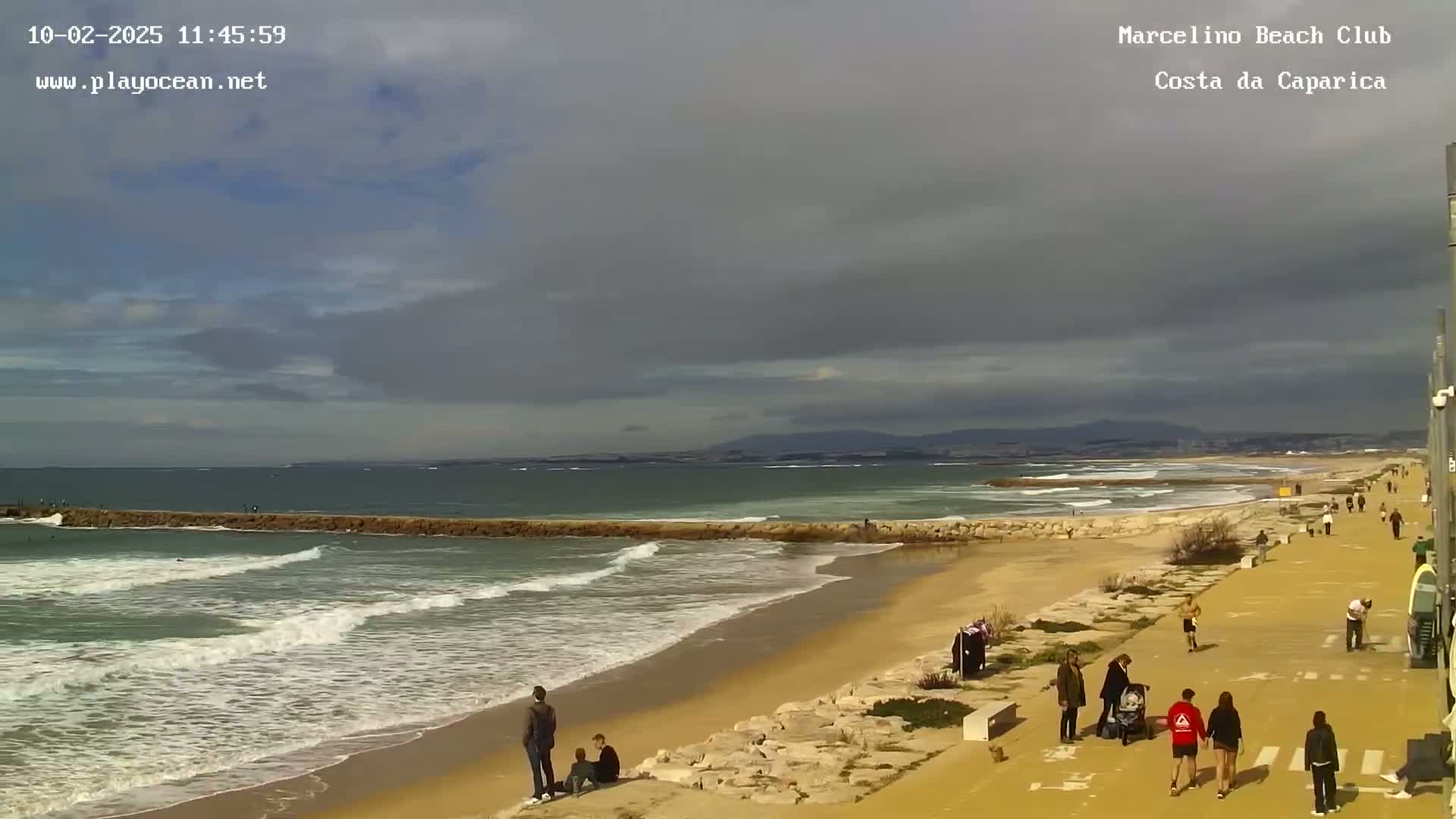 A sandy beach with people walking along a paved path, waves breaking on the shore under a partly cloudy sky.